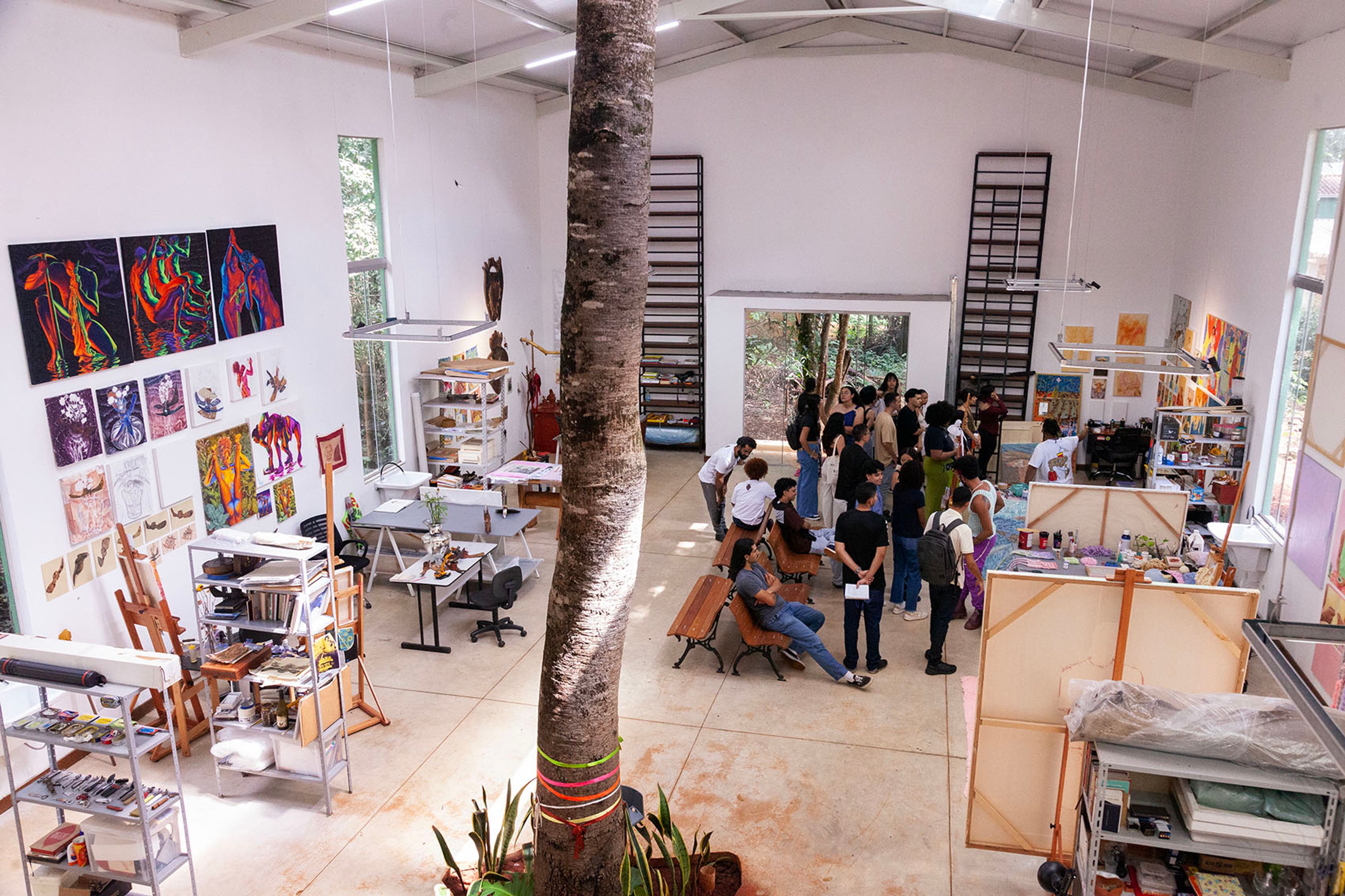 A bright art studio with a large tree trunk in the center, diverse artworks on the walls, and a group of people gathered.