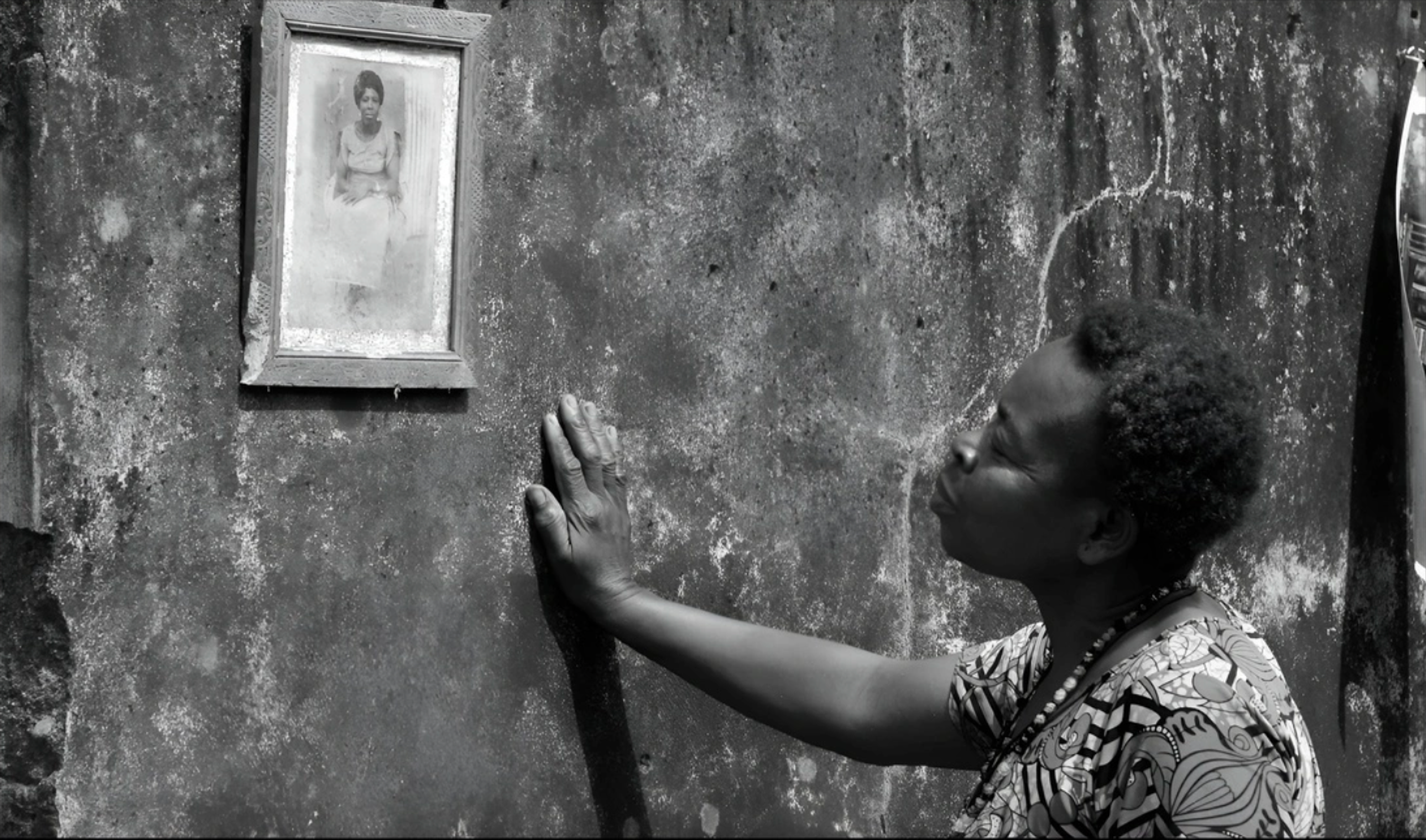 A black and white photo shows a woman puckering her lips to kiss a textured wall below a framed portrait of another woman.