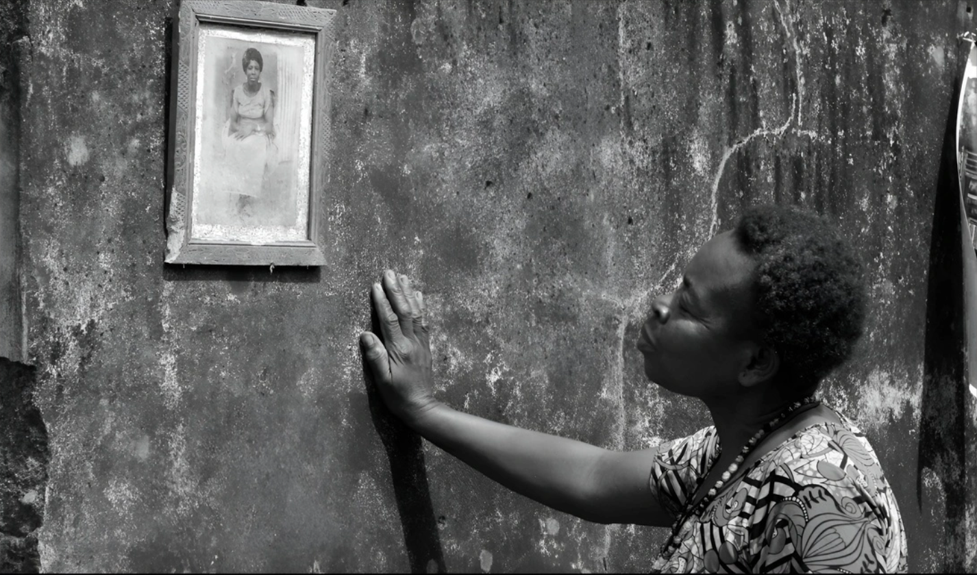 A black and white photo shows a woman puckering her lips to kiss a textured wall below a framed portrait of another woman.
