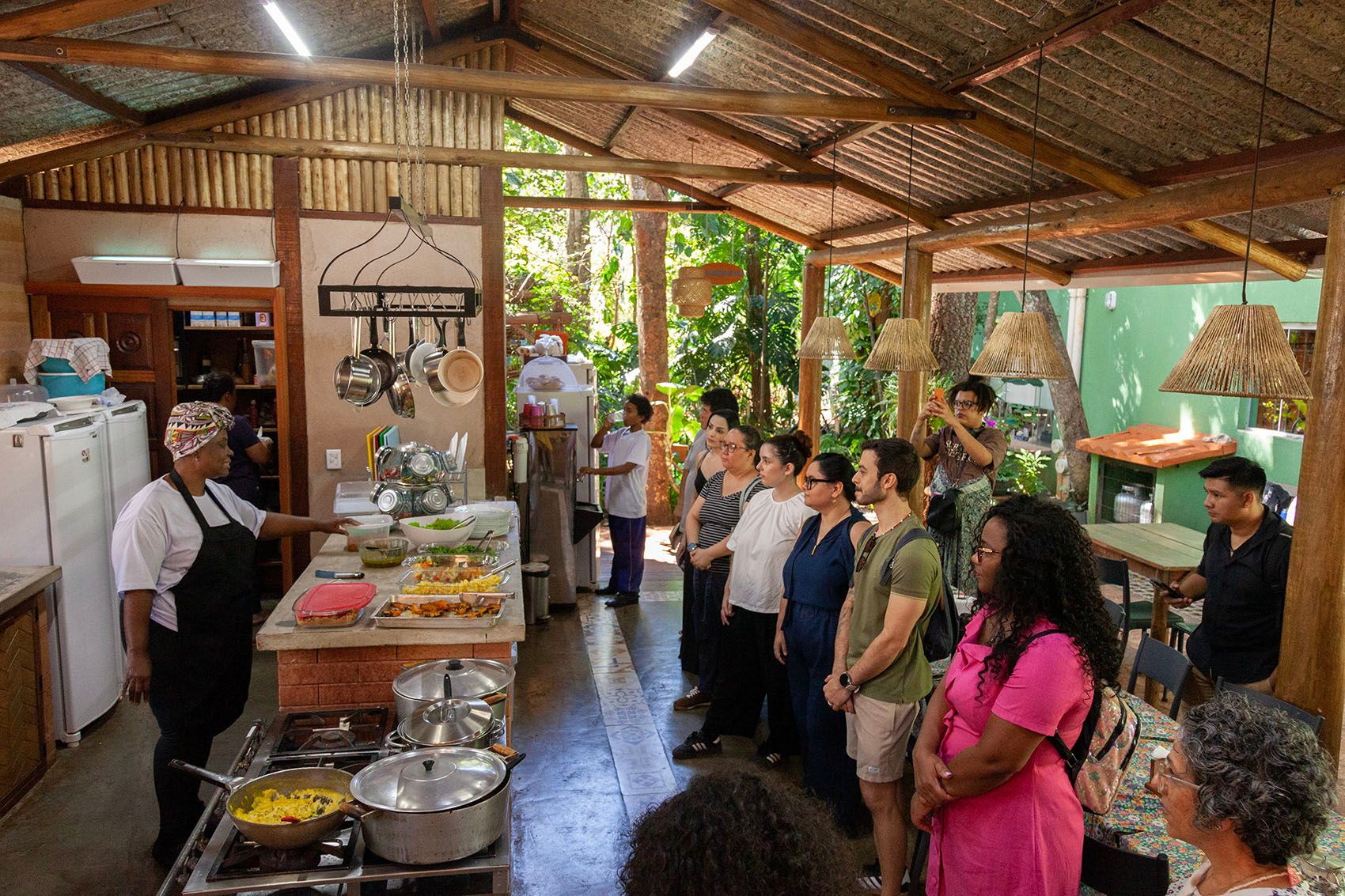 A chef presents food on a counter to a group of people in a rustic, open-air kitchen.
