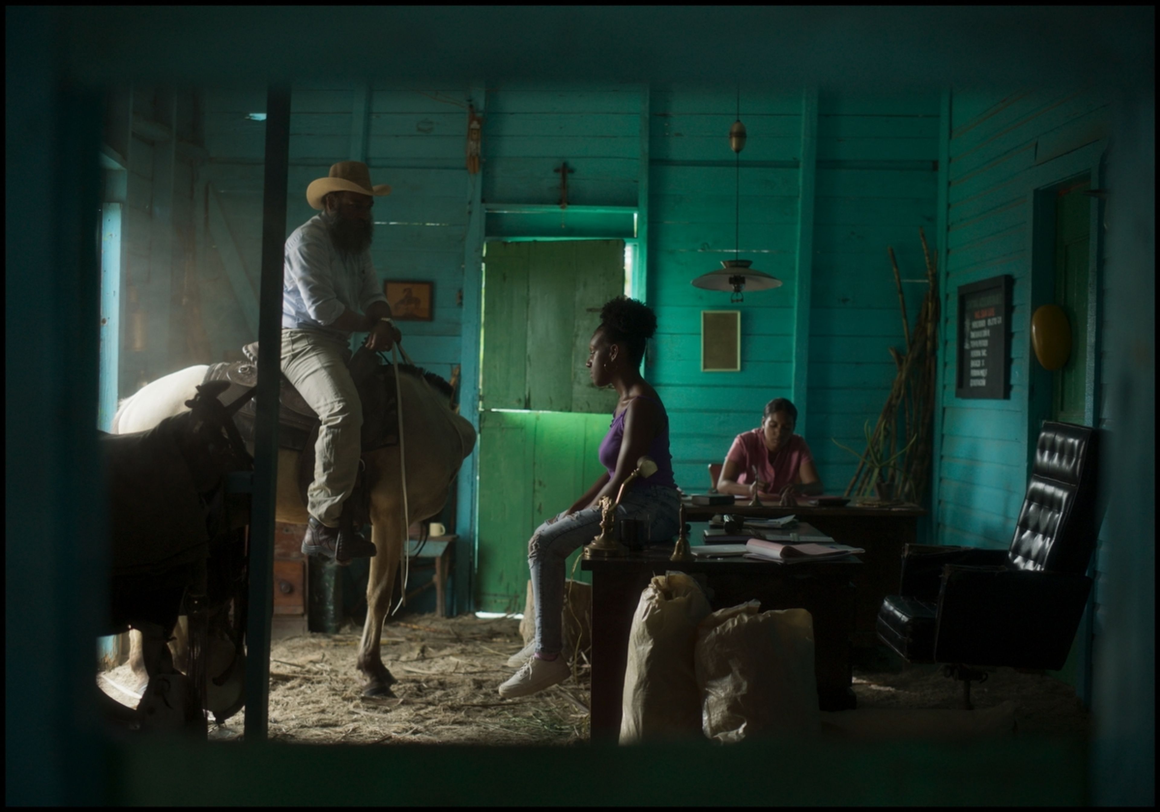 A man on horseback inside a rustic teal office speaks with a woman, another person is at a desk.