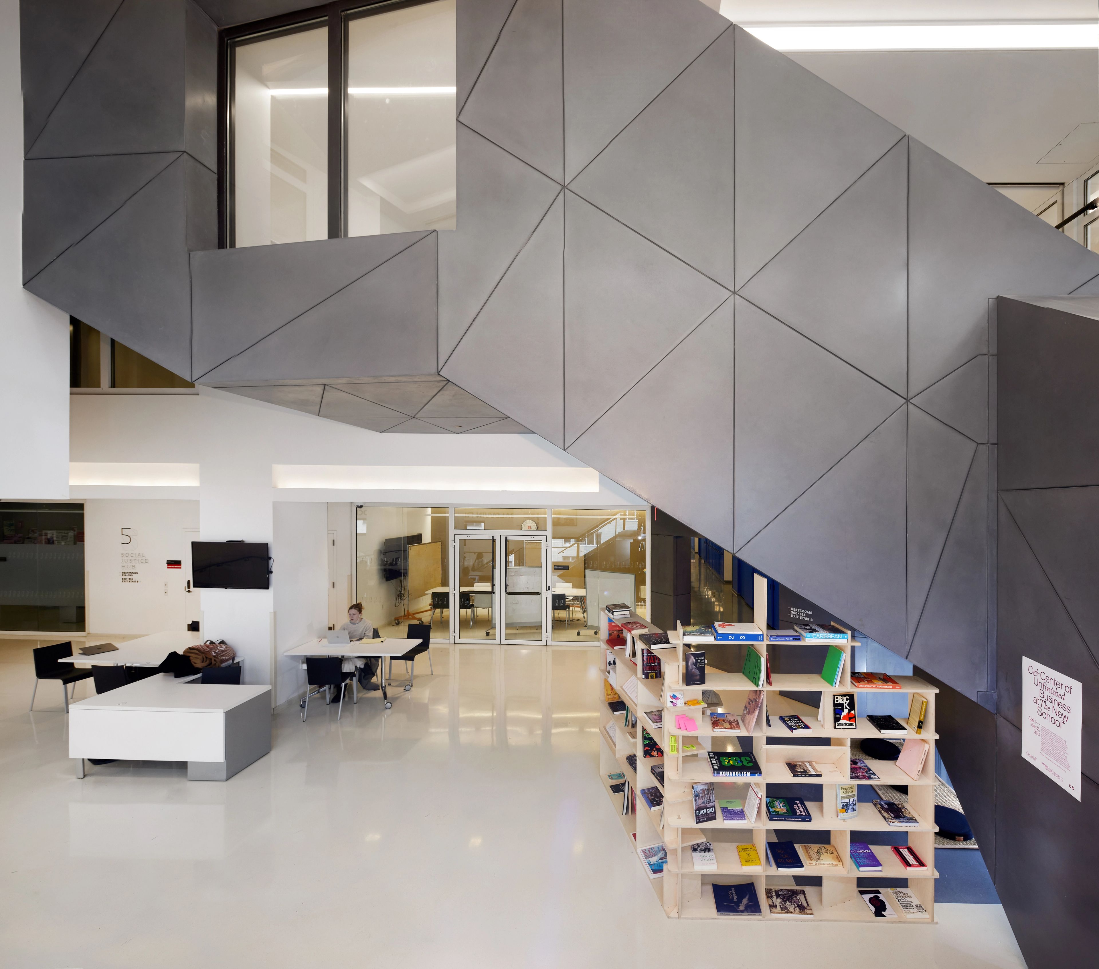 Modern interior featuring a geometric gray paneled staircase, a modular bookshelf, and a person working at a table.