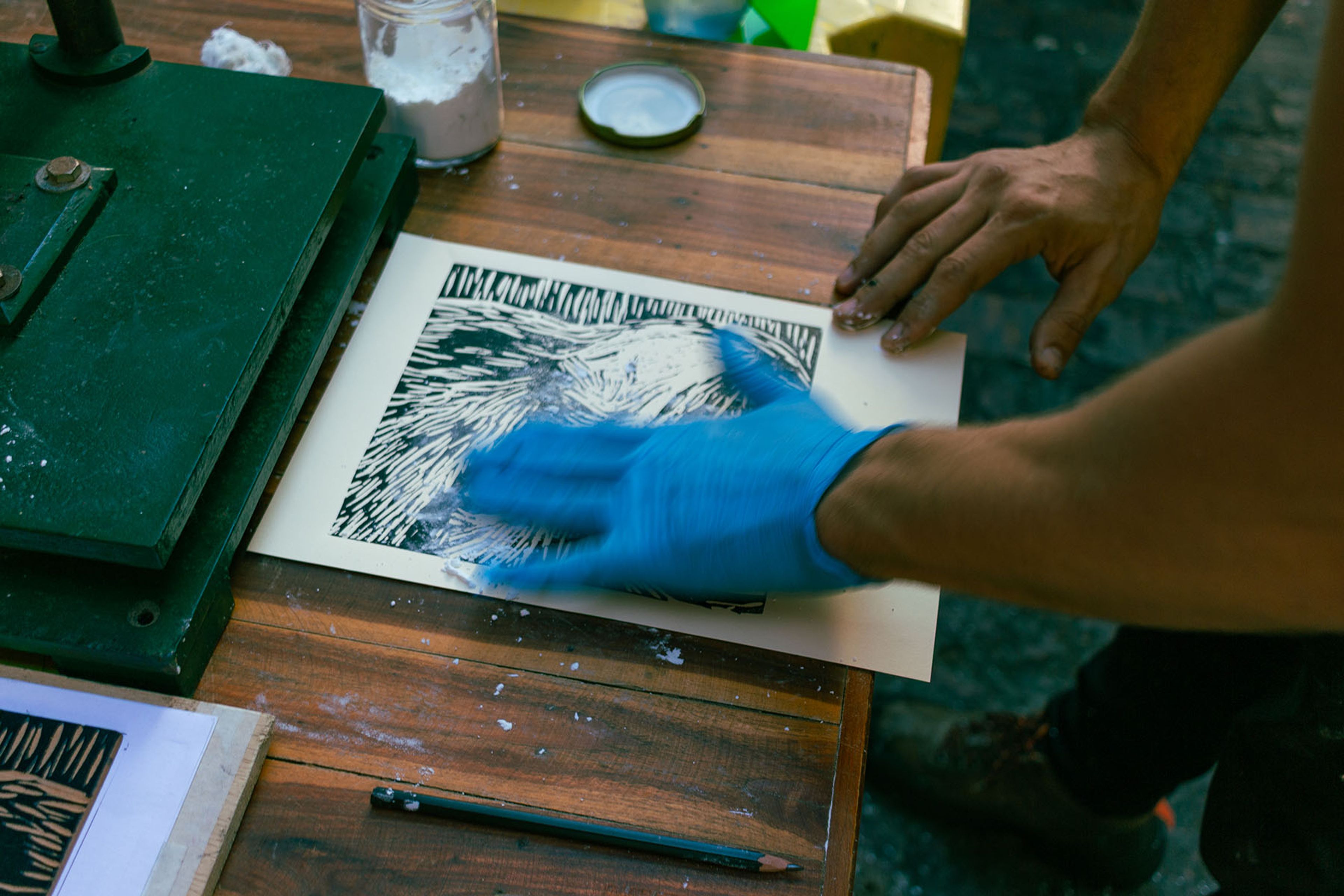 A person in a blue glove rubs a black and white print on a wooden table next to a green printing press.