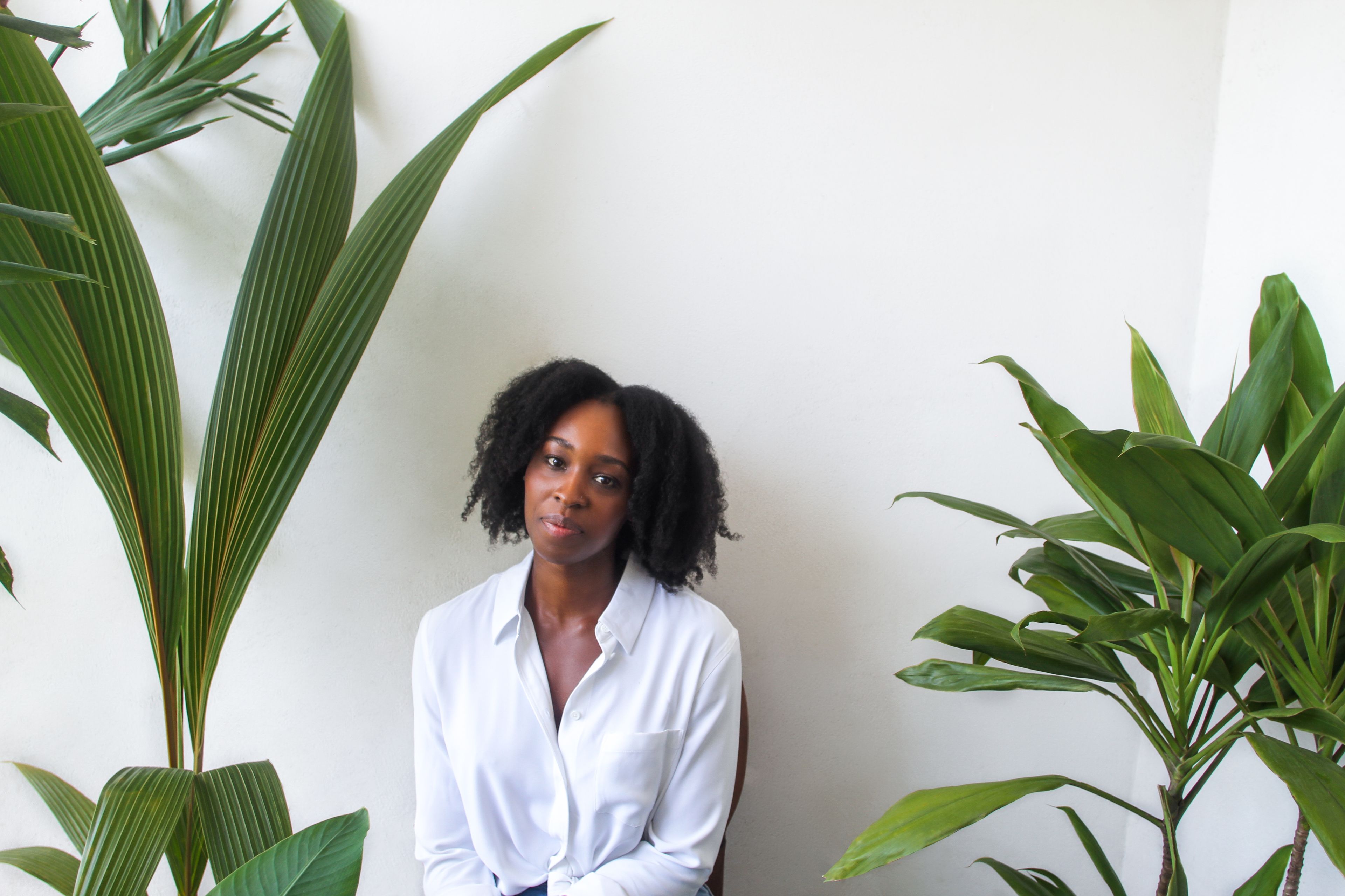 A Black woman in a white shirt sits between two large green plants against a white wall.