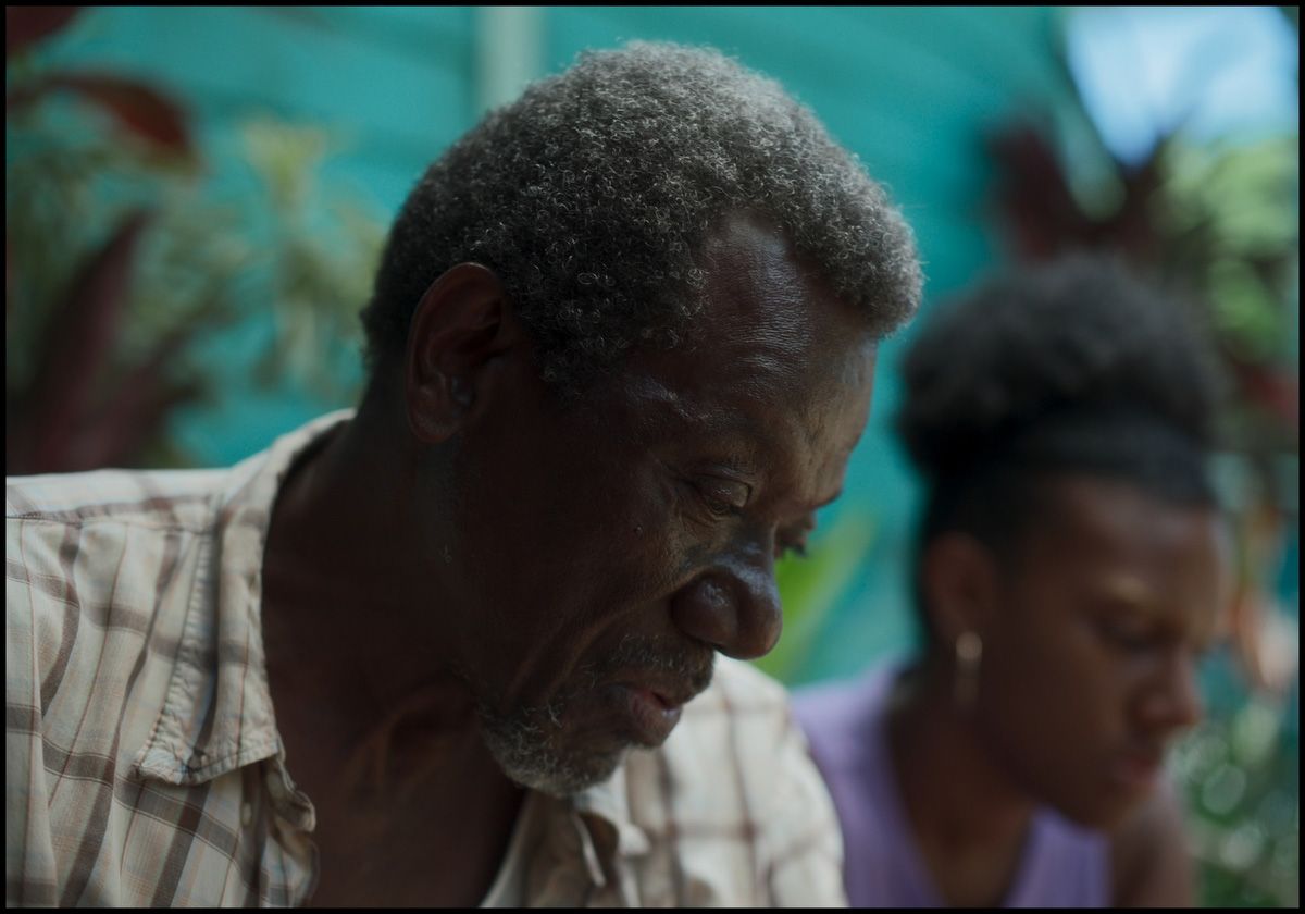 An older Black man in a plaid shirt looks down, with a younger Black woman blurred in the background also looking down.