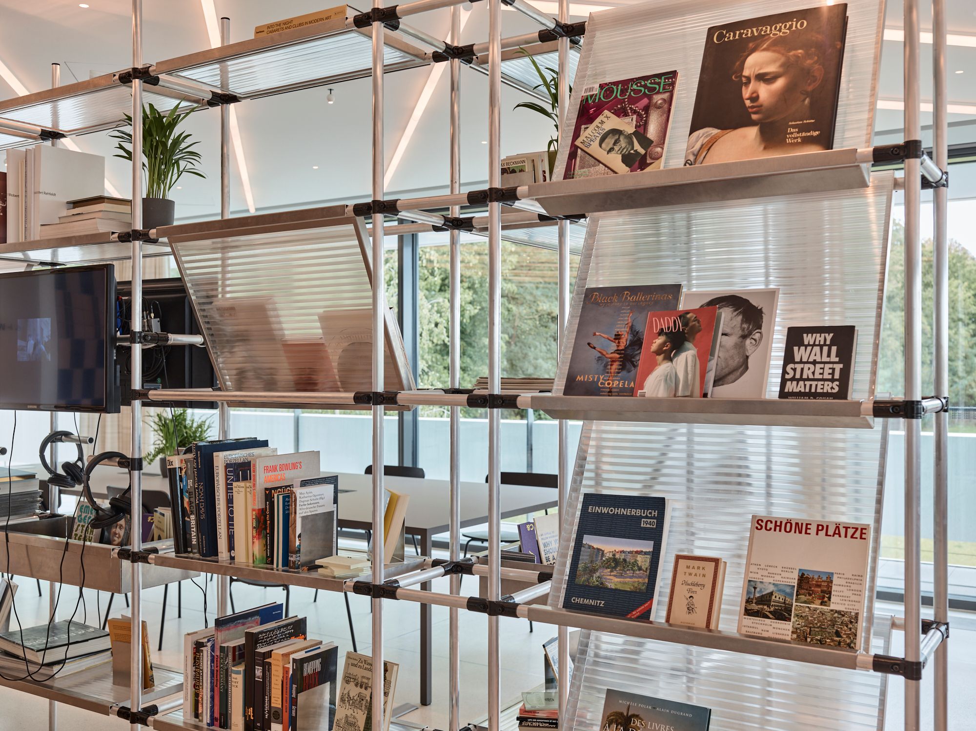 A modern modular bookshelf made of metal pipes, displaying books, magazines, and a TV, with large windows in the background.