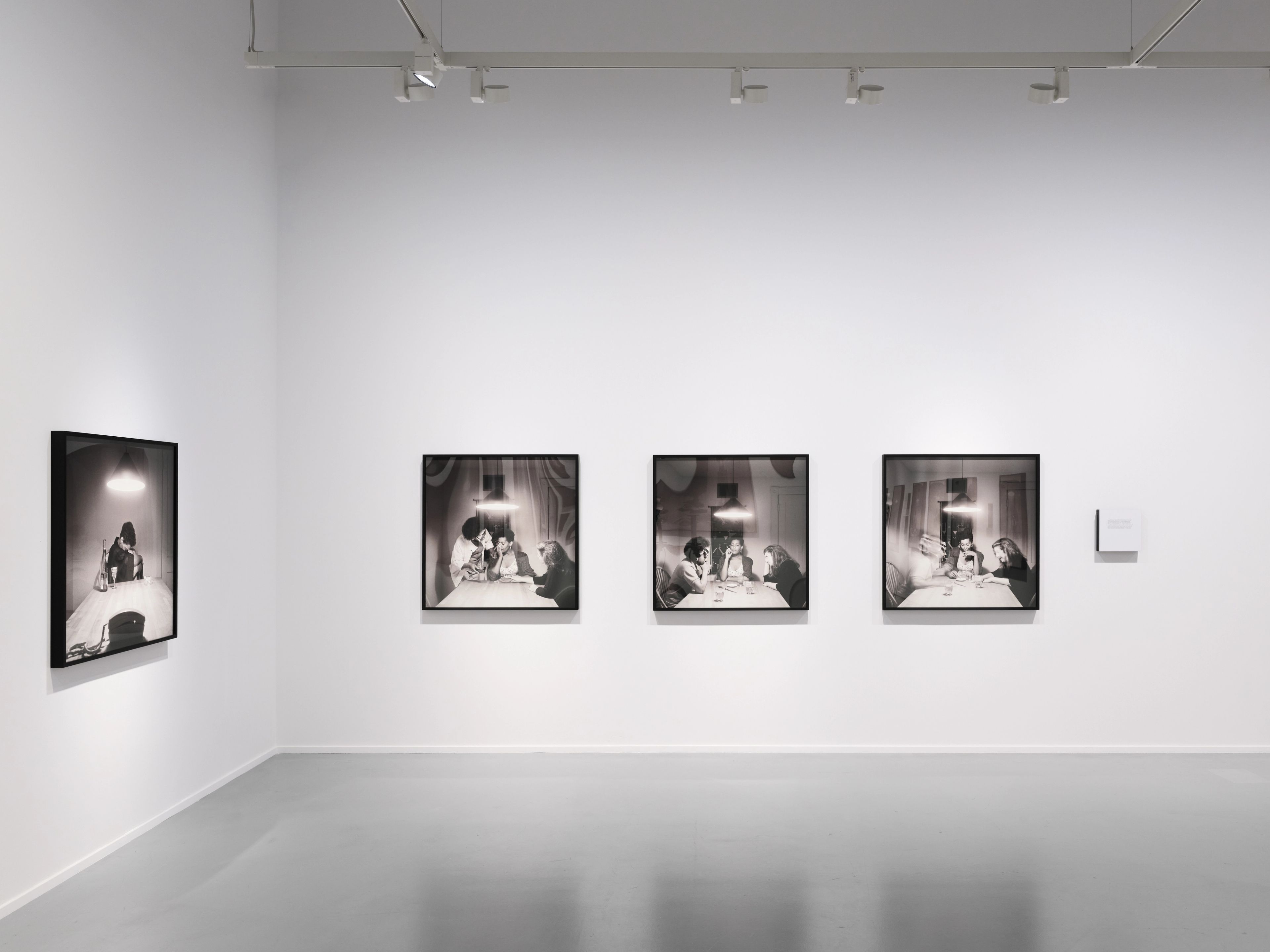 Four framed black and white photographs, depicting figures at a table under a lamp, displayed in a white-walled gallery.