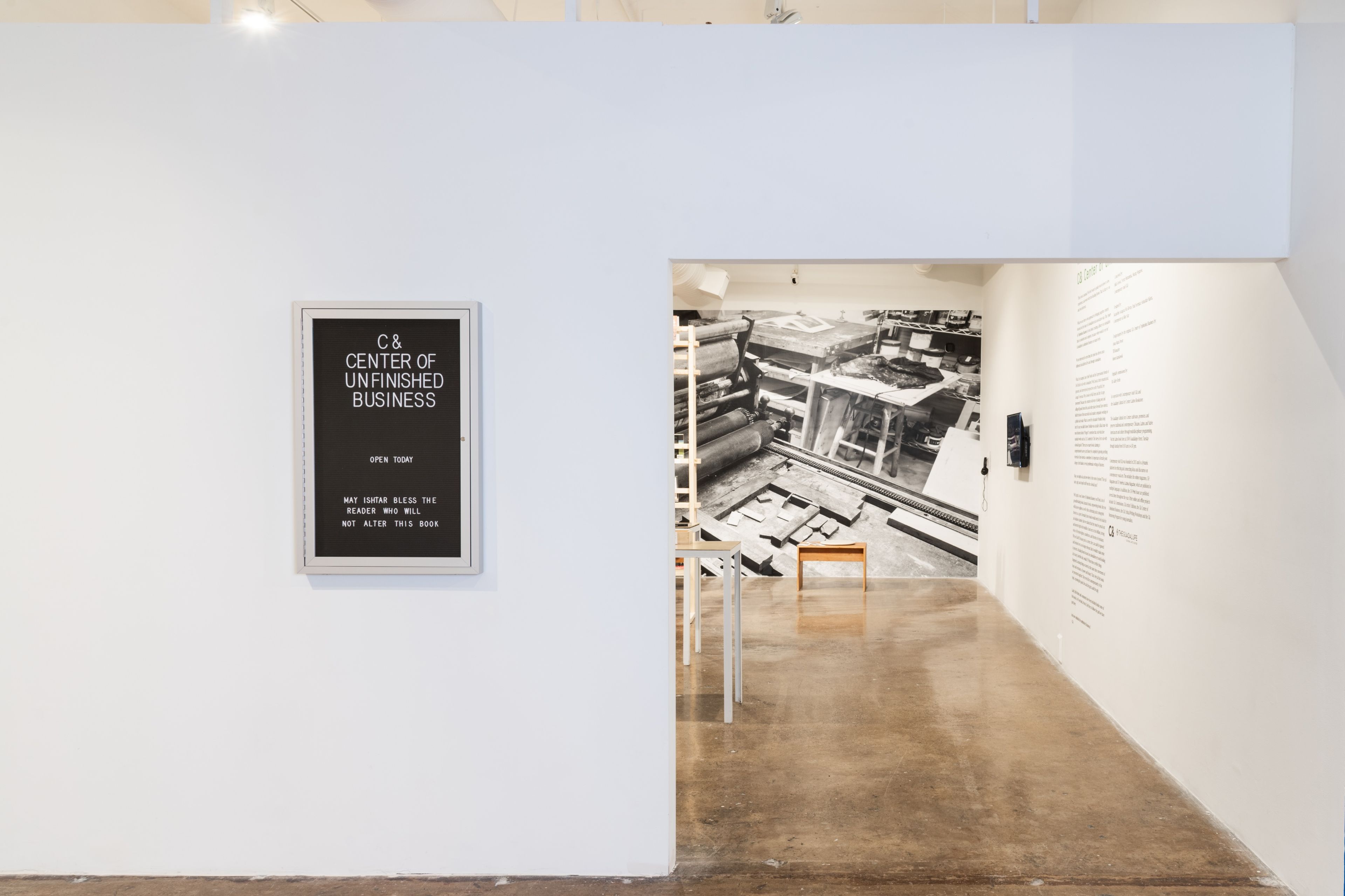 A white wall features a black sign for "C & Center of Unfinished Business" next to an open doorway leading into a room with a large black and white mural of a workshop and explanatory text on the right wall.