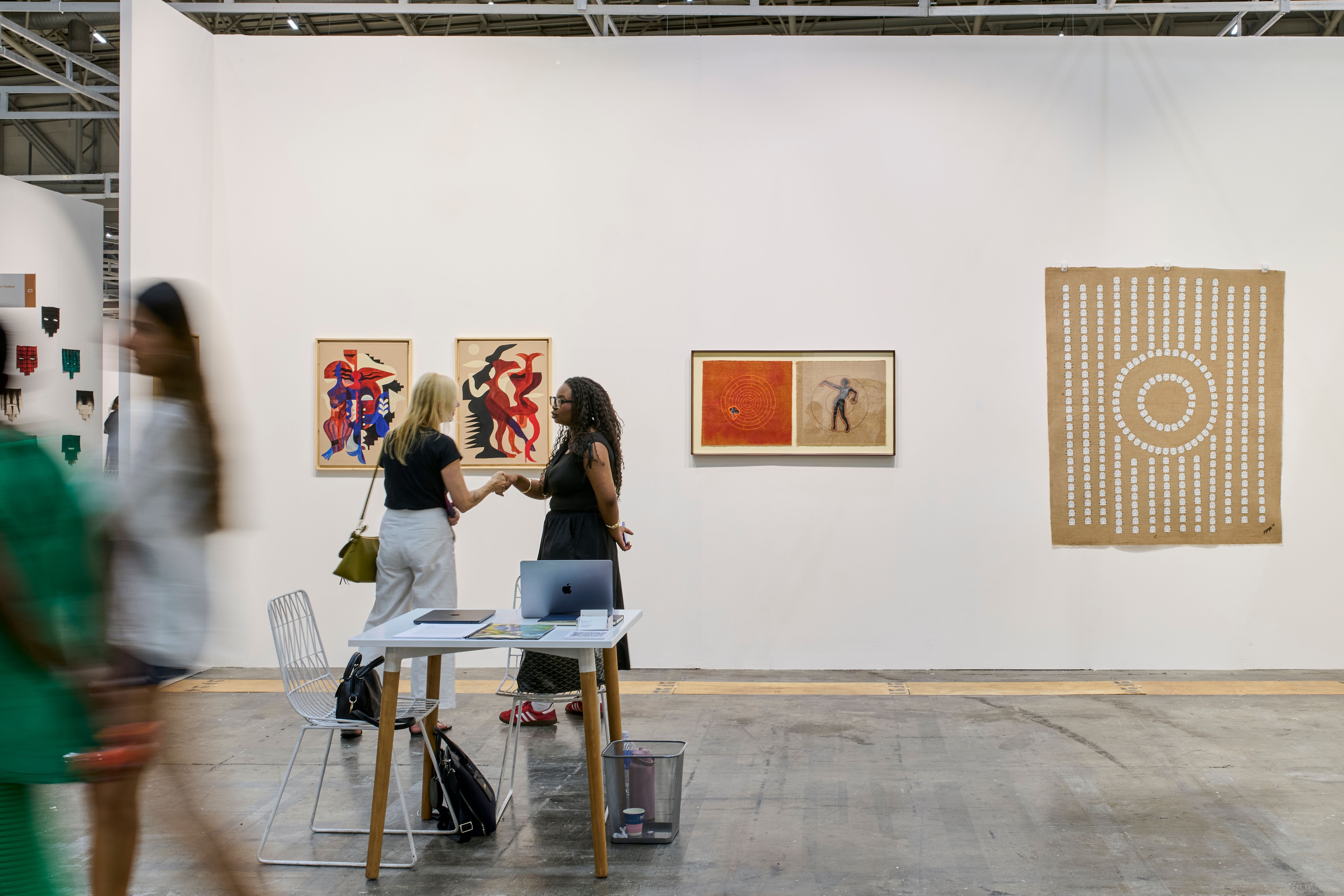 An art gallery booth with diverse artworks on white walls, two women conversing at a table, and blurred figures walking in the foreground.