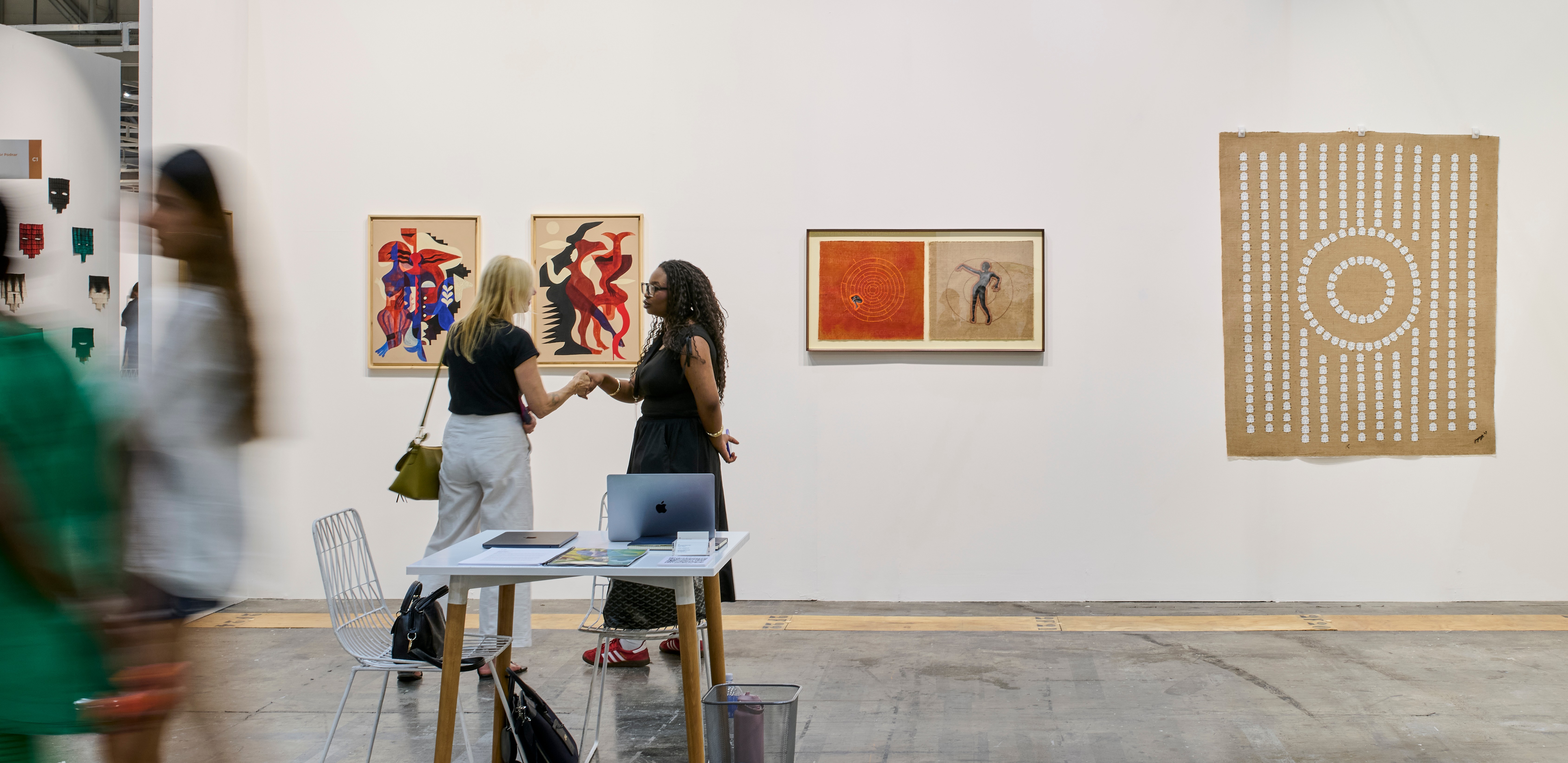 An art gallery booth with diverse artworks on white walls, two women conversing at a table, and blurred figures walking in the foreground.