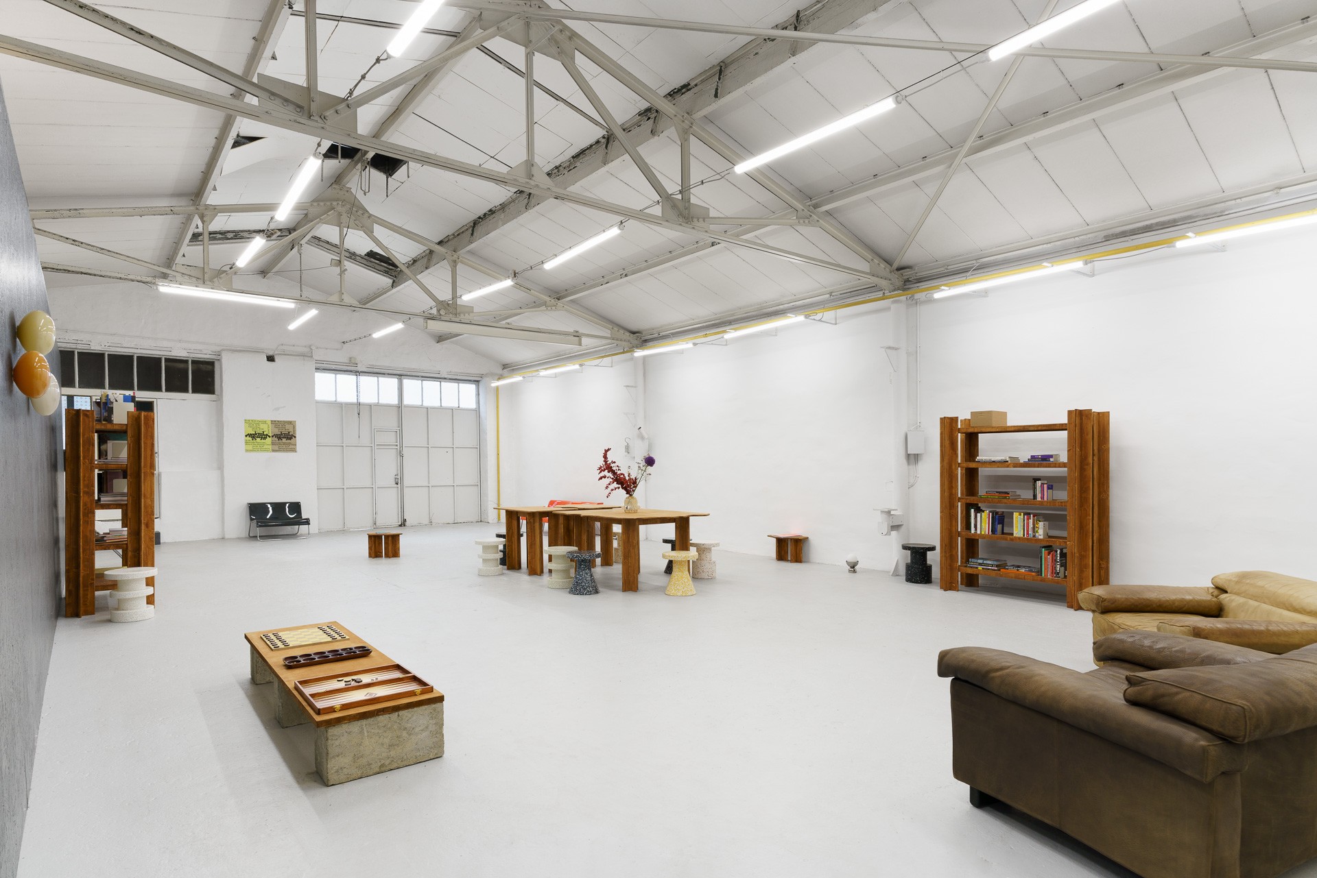 A spacious, brightly lit white room with an industrial ceiling, sparsely furnished with wooden tables, bookshelves, colorful stools, and leather seating, including a backgammon game.