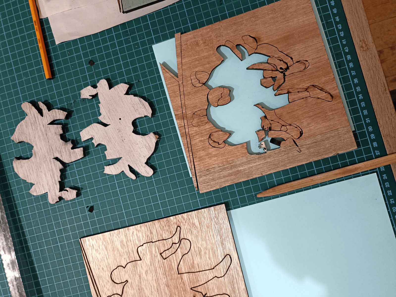 Intricate wooden character shapes, some cut out to reveal a blue layer, are laid on a green cutting mat.