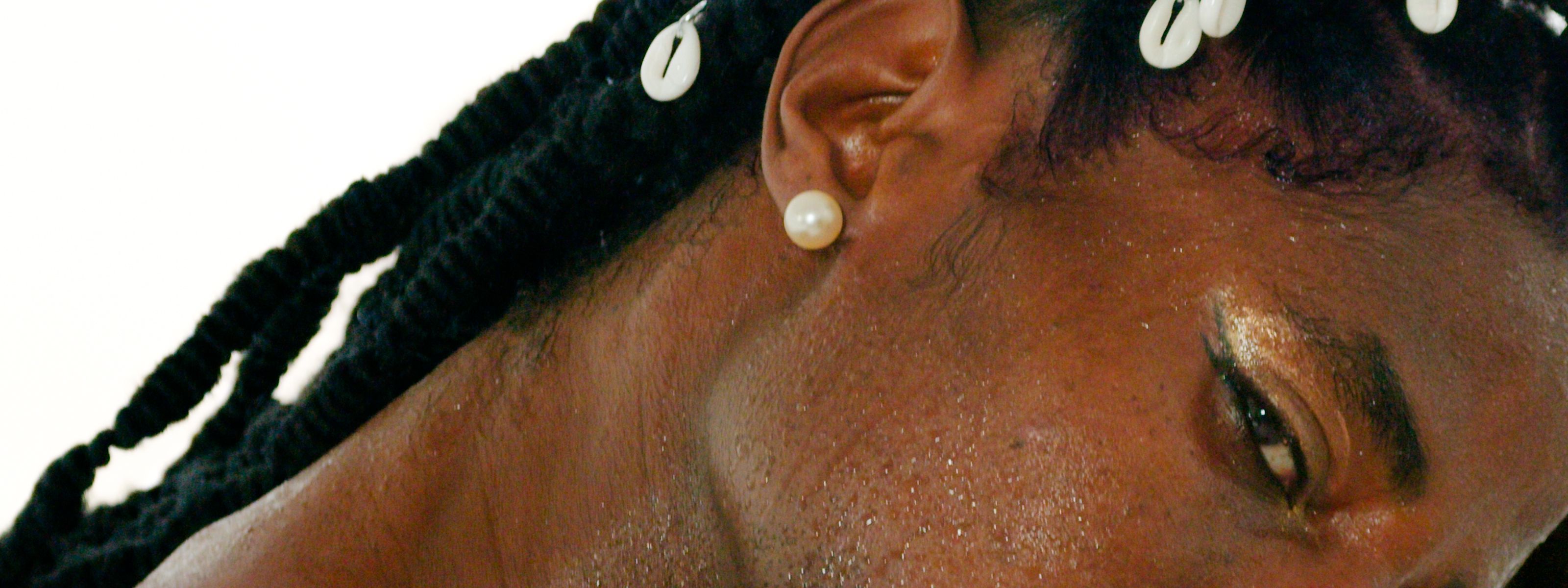 Close-up of a person's face with dark skin, black dreadlocks with cowrie shells, a pearl earring, and gold eyeshadow.