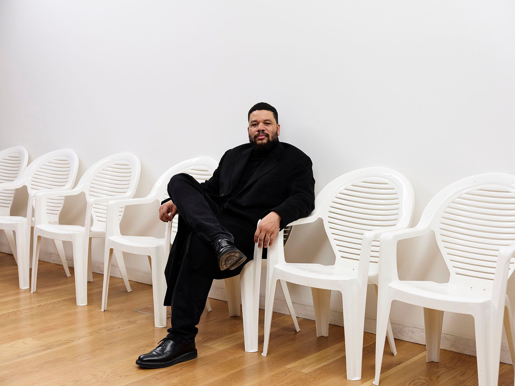 A man in a black coat sits with crossed legs in a white plastic chair, part of a row against a white wall.