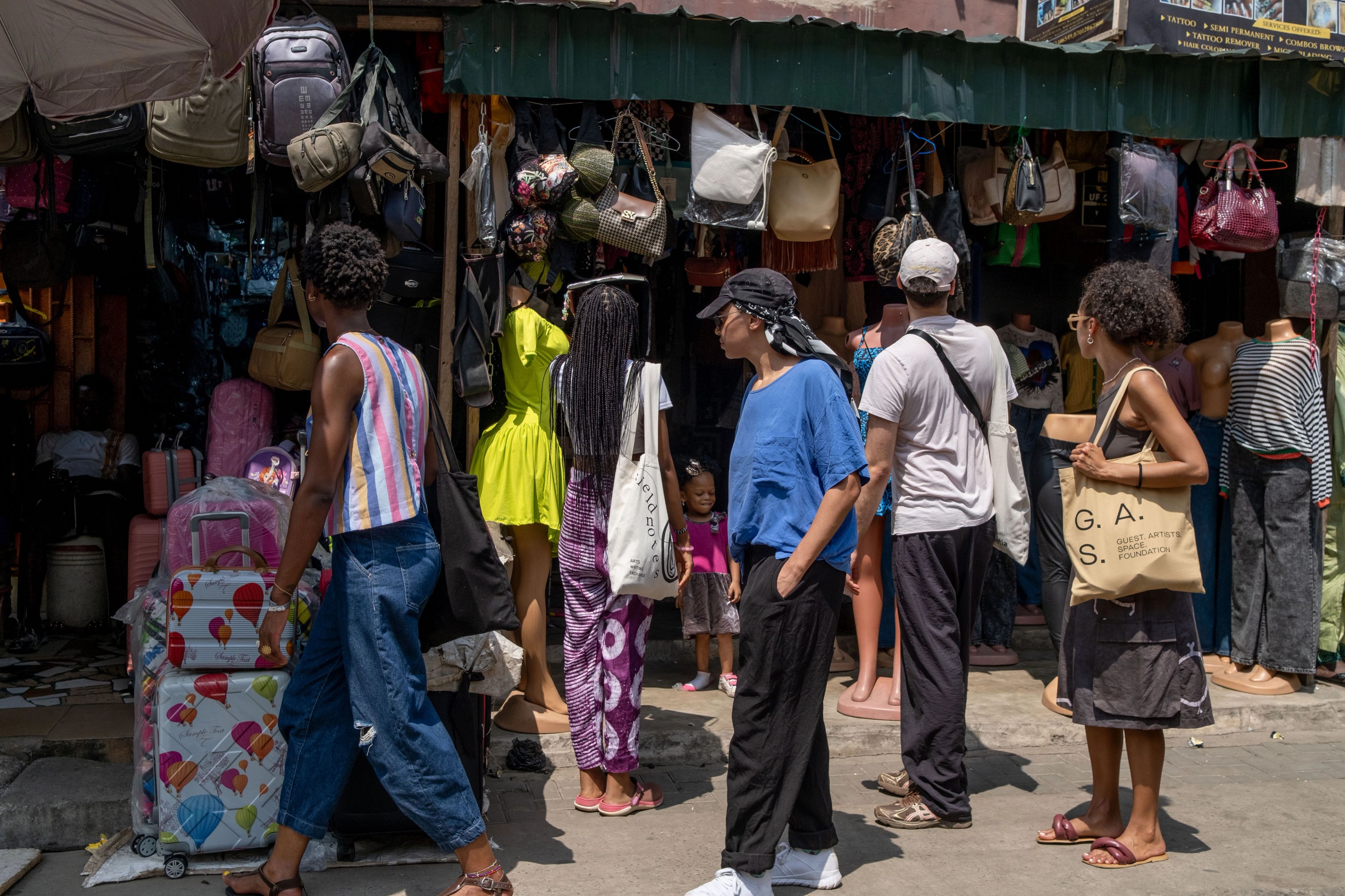 Several people walk past a street market stall displaying bags, luggage, and clothing.