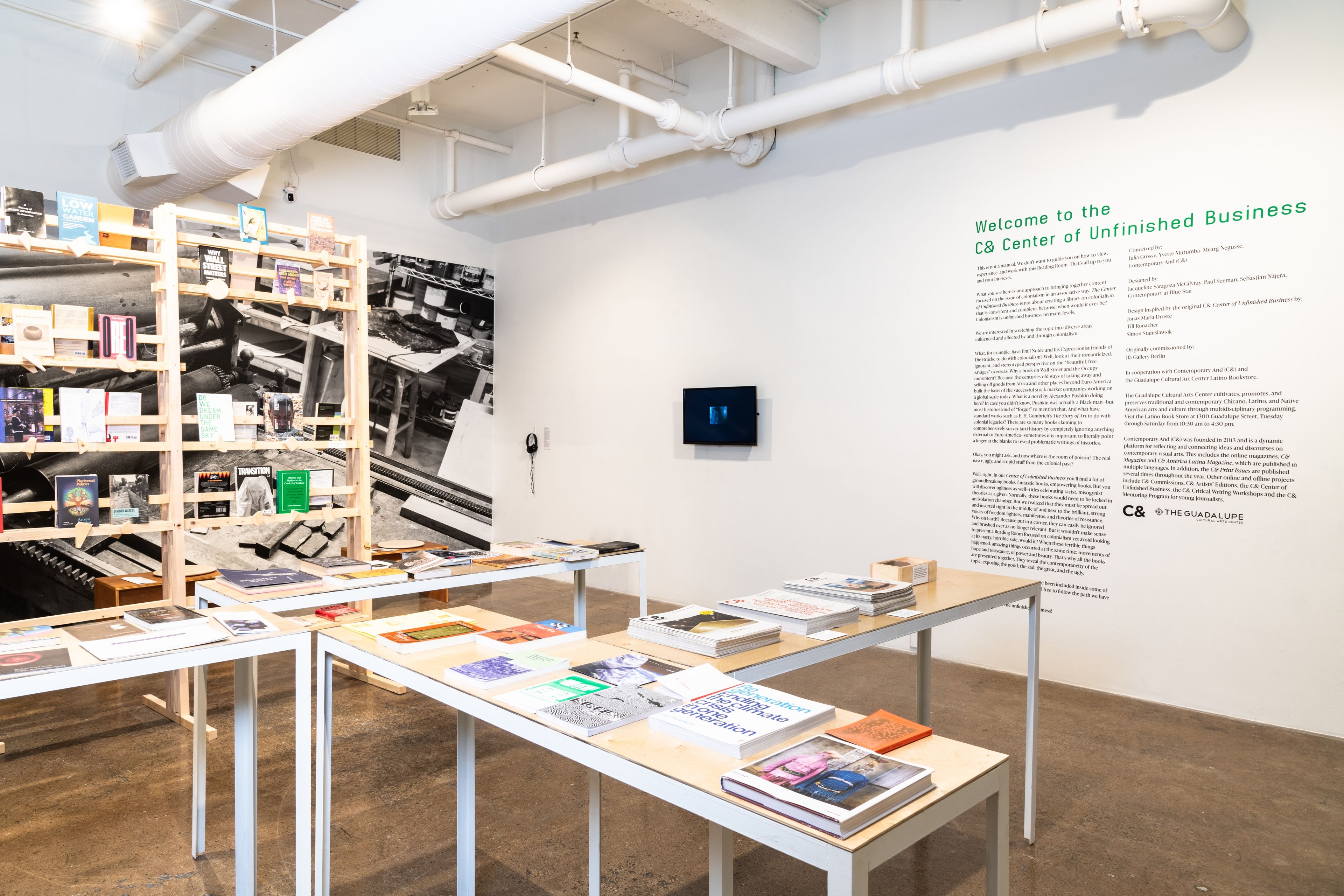 Exhibition space with publications displayed on light wooden tables and shelves, a large black and white photo mural, and wall text "Welcome to the C& Center of Unfinished Business".