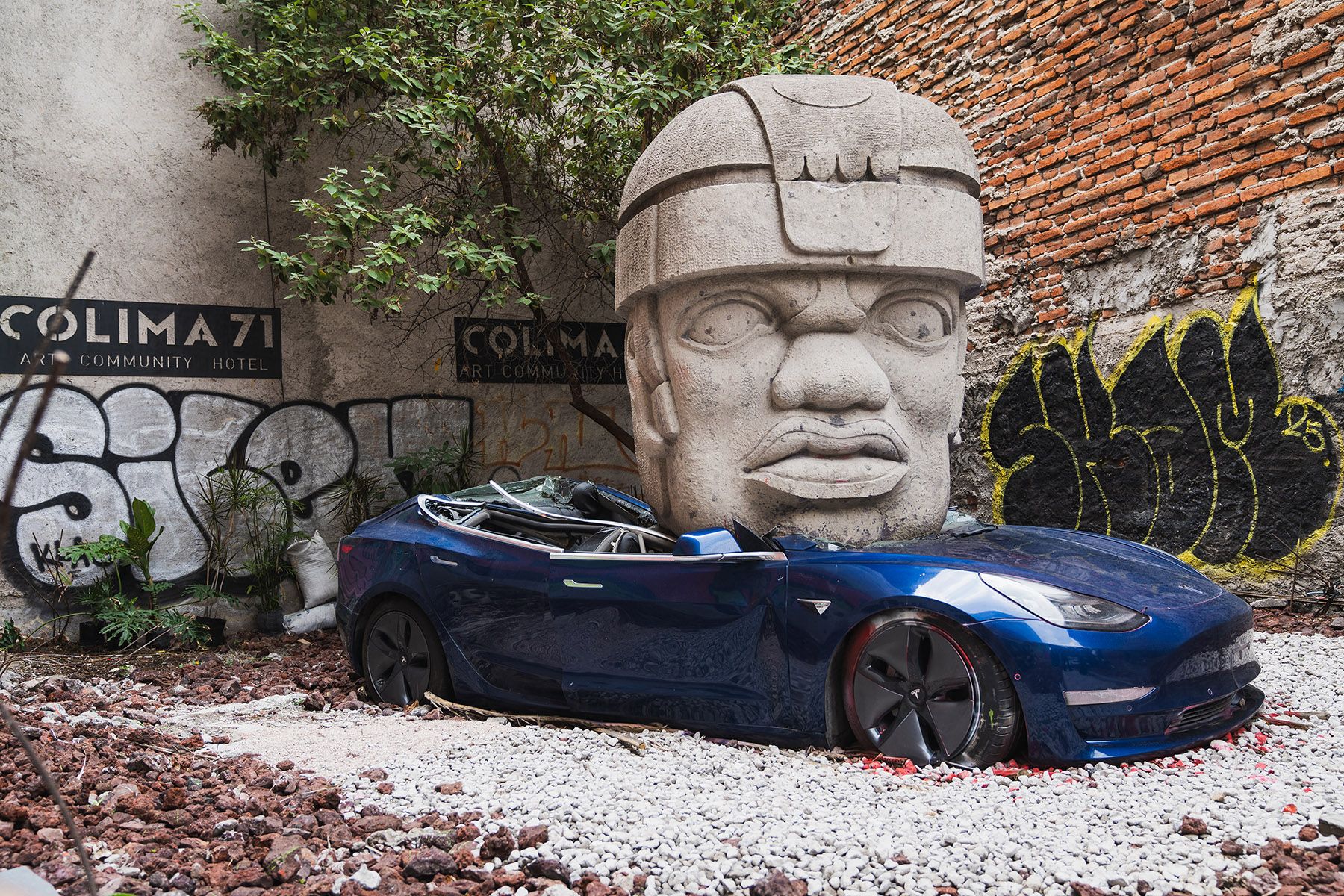 A large stone Olmec head sculpture emerges from a blue Tesla car, with graffiti-covered walls in the background.