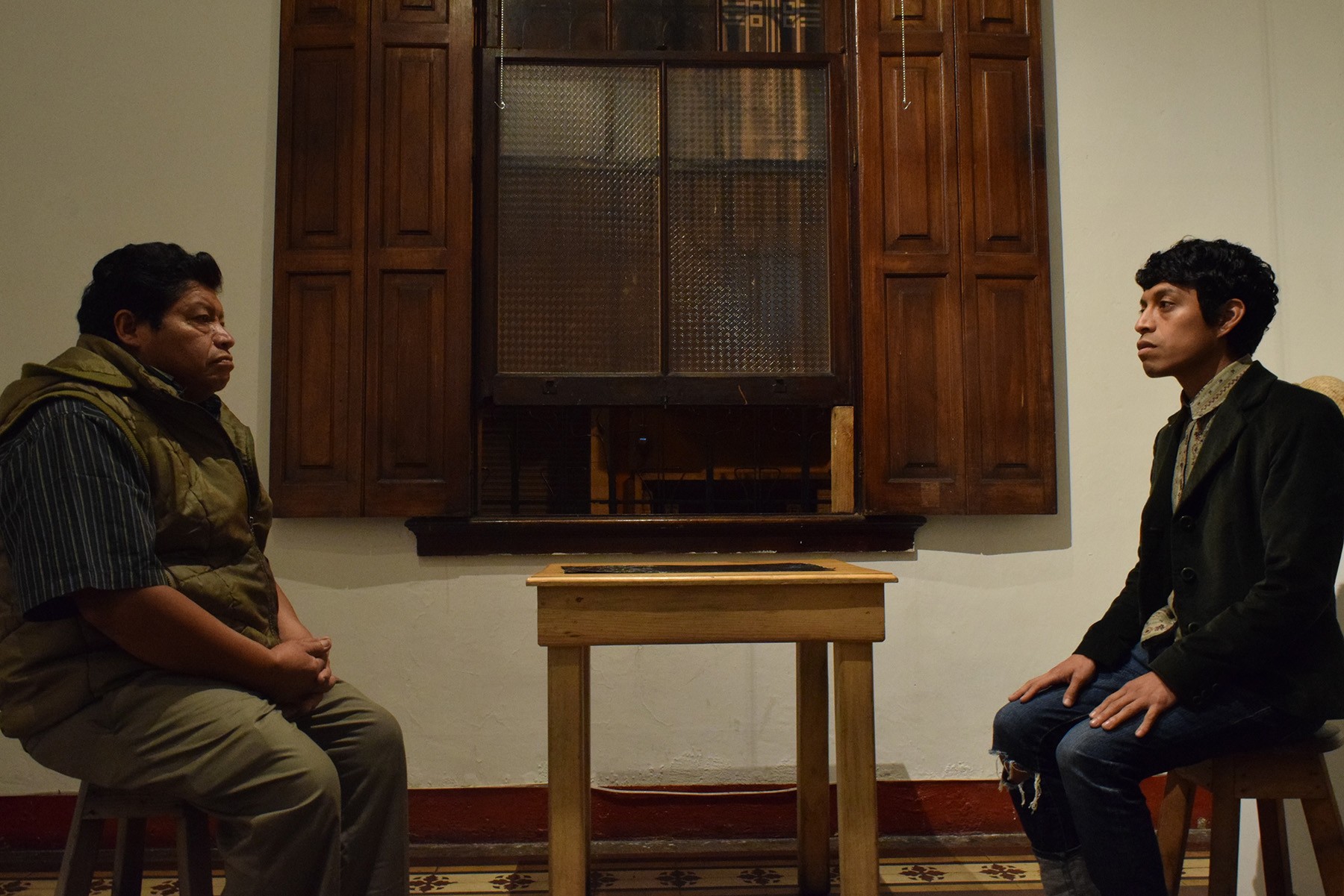 Two men sit on opposite sides of a small table, each looking forward, with a large window and wooden shutters behind them.