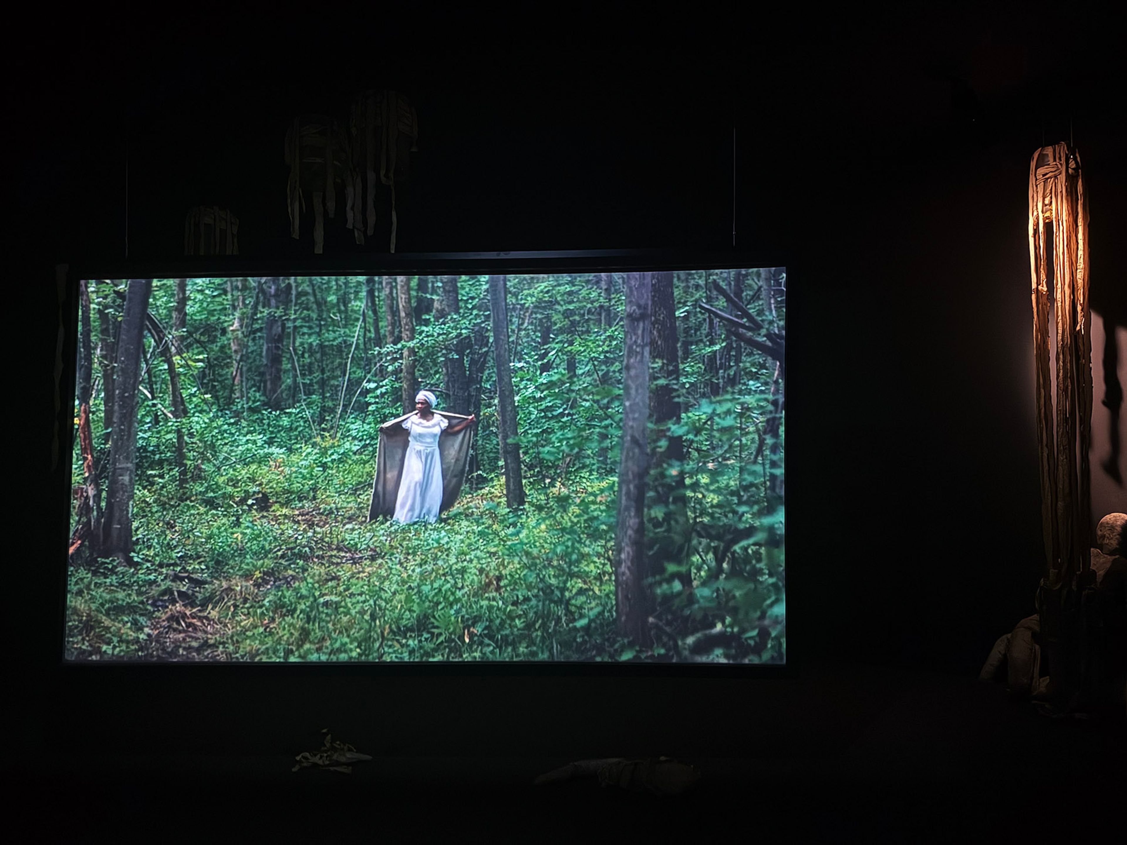 A dark room features a large projection of a woman in a white dress in a lush forest, with a tall, illuminated sculpture on the right.