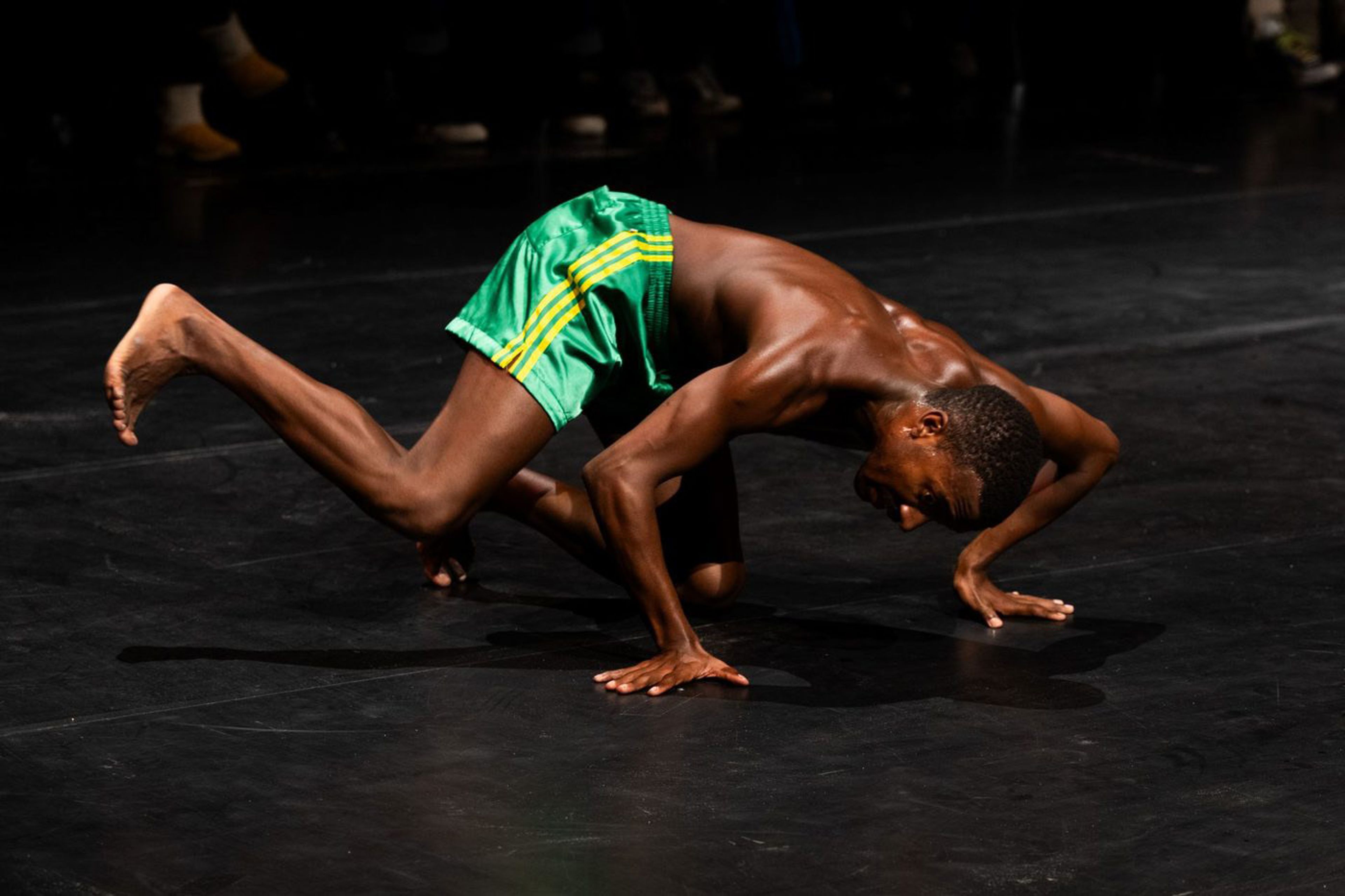 A dancer in green shorts is in a low, crouched pose on a dark stage.