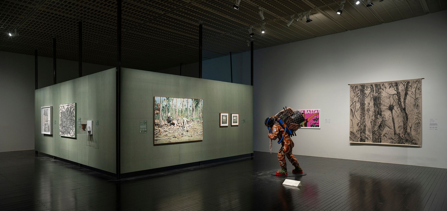 Art gallery featuring a person-like figure in an orange patterned suit with a huge backpack, surrounded by various artworks on walls.