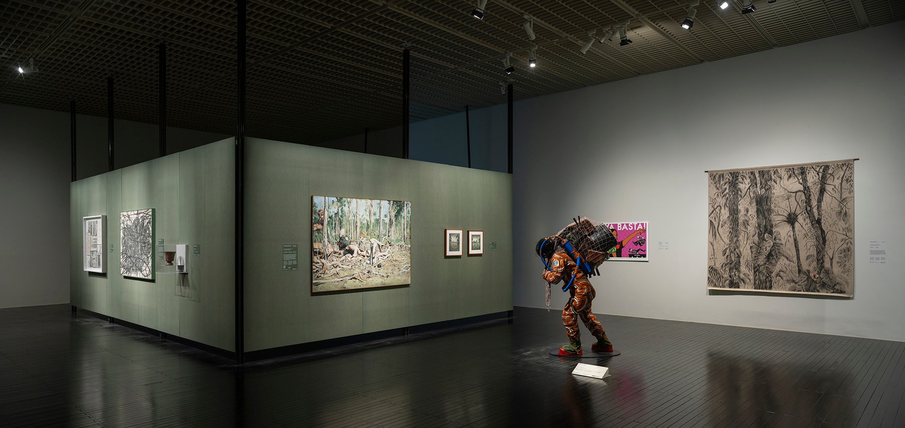 Art gallery featuring a person-like figure in an orange patterned suit with a huge backpack, surrounded by various artworks on walls.