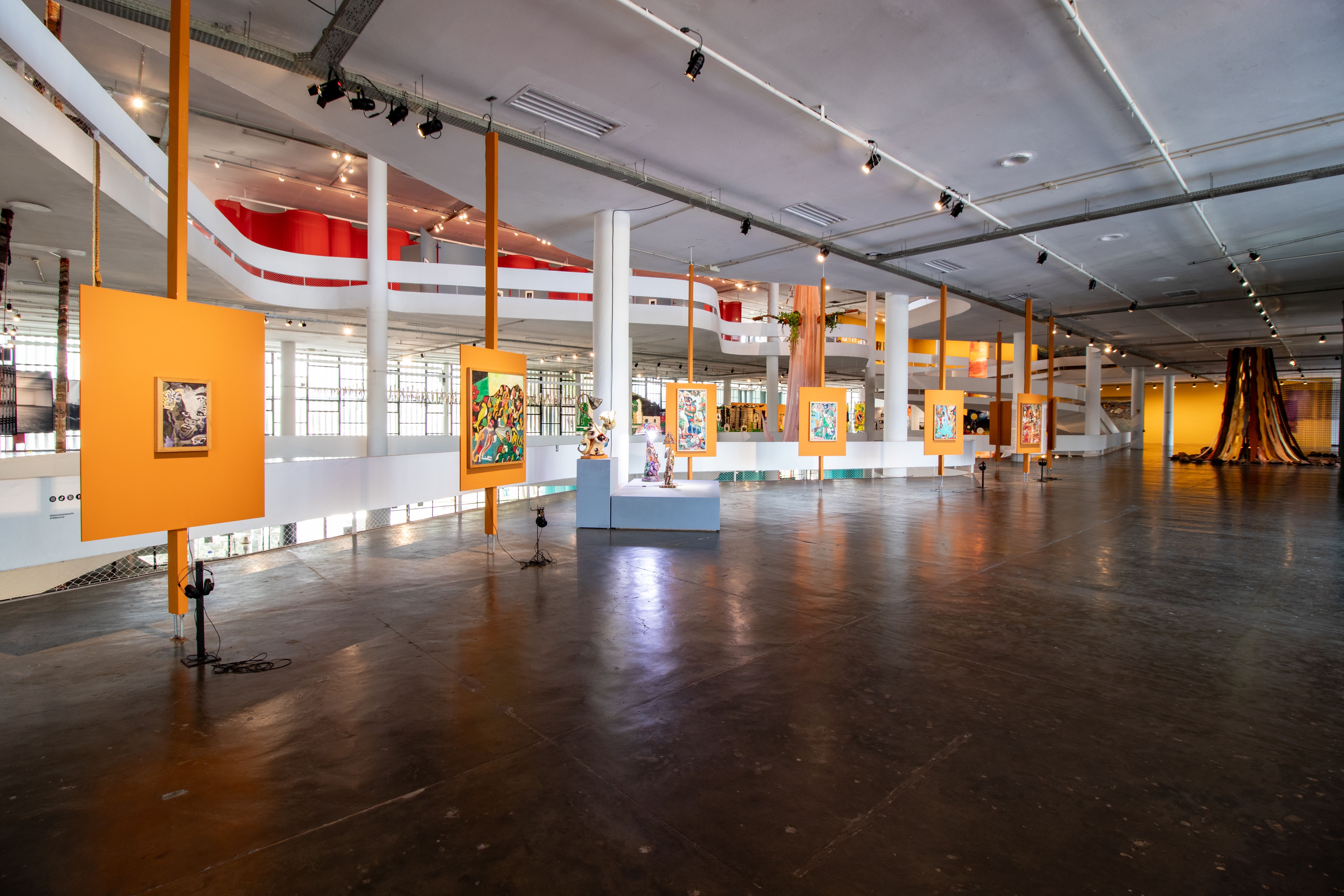 A large, brightly lit art gallery with paintings on suspended orange panels, an upper level with a red railing, and a golden fabric sculpture in the background.