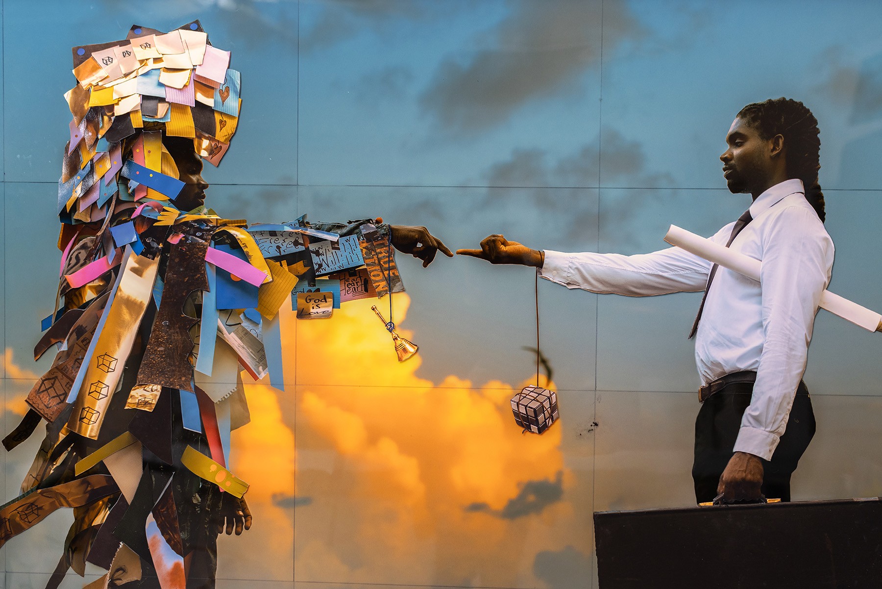 Two figures recreate 'The Creation of Adam' against a dramatic sky. The left figure wears a vibrant, multi-material costume; the right is a man in a business shirt holding rolled papers, a small cube dangling from his finger.