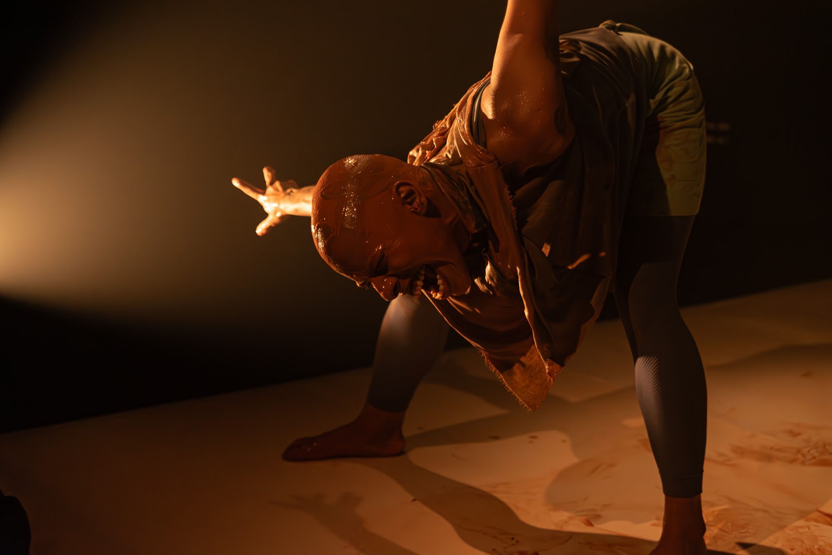 A person covered in reddish-brown mud leans forward dramatically with an outstretched arm and an intense expression, lit by a spotlight.