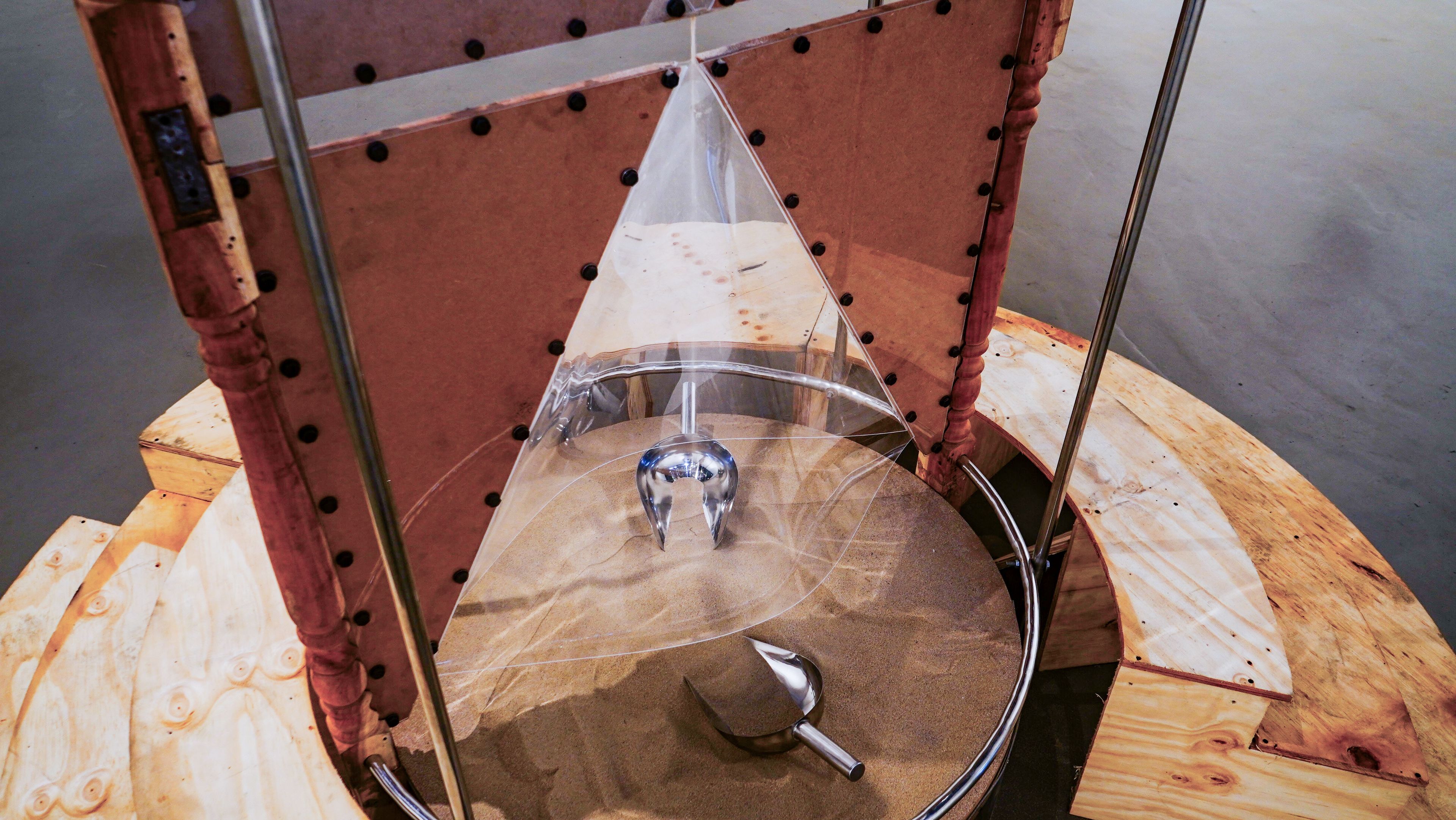 A Foucault pendulum swings over a circular sand bed, creating patterns, with a metal scoop in the sand.