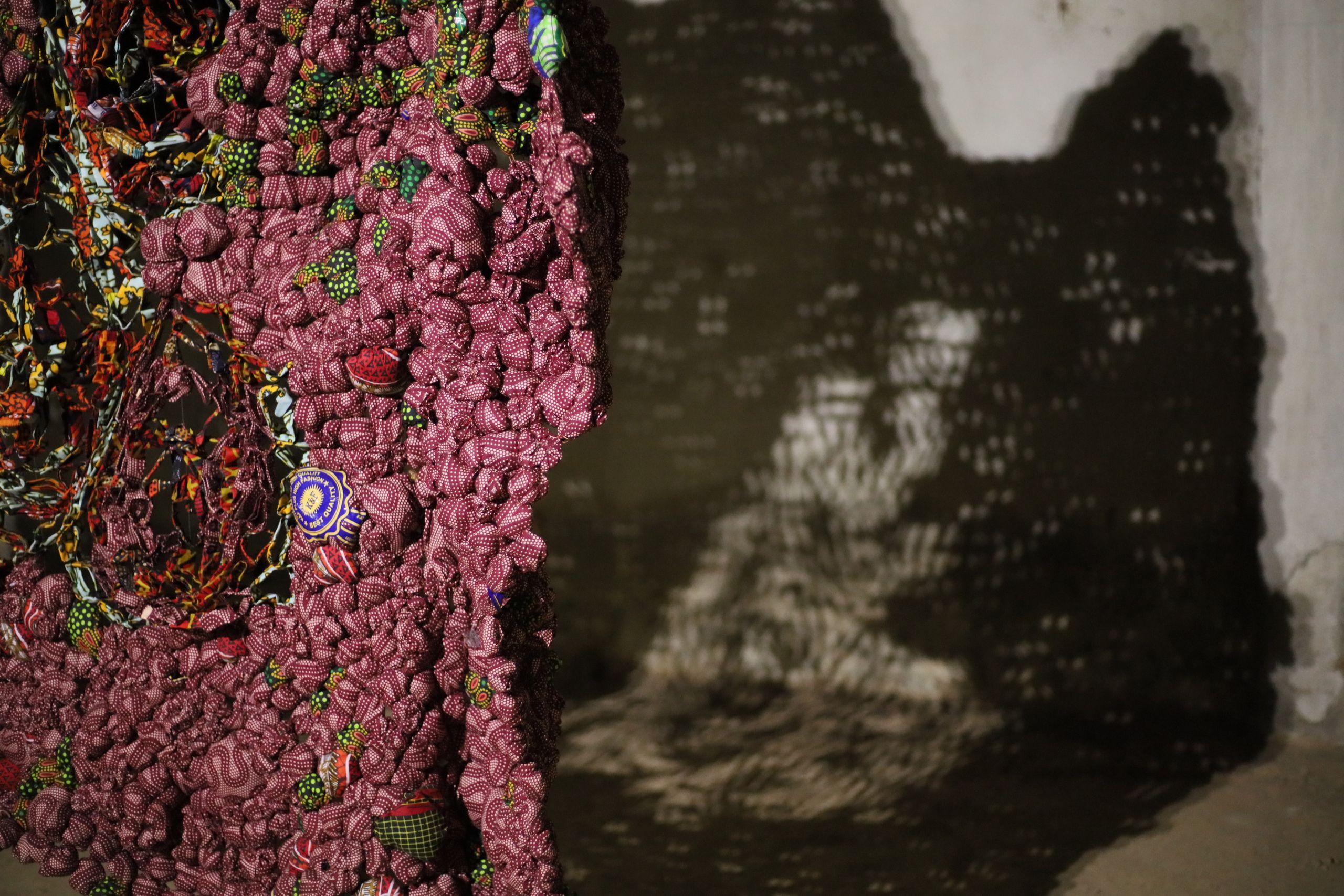 Lumpy reddish-purple textile art with colorful details, casting a dappled shadow on a wall.