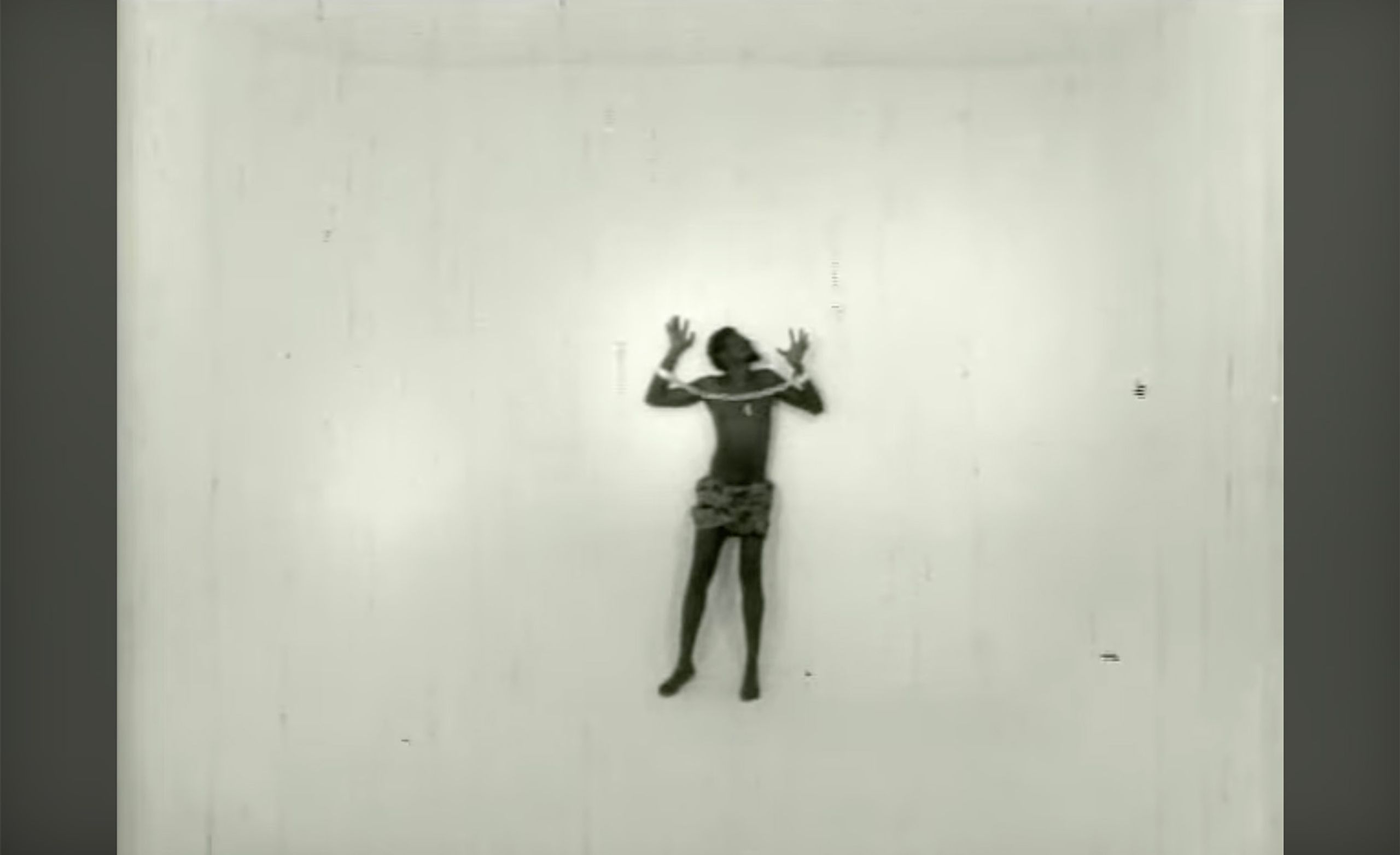 A dark-skinned person, hands raised and bound by ropes across their shoulders, wearing a patterned skirt, against a plain white wall.