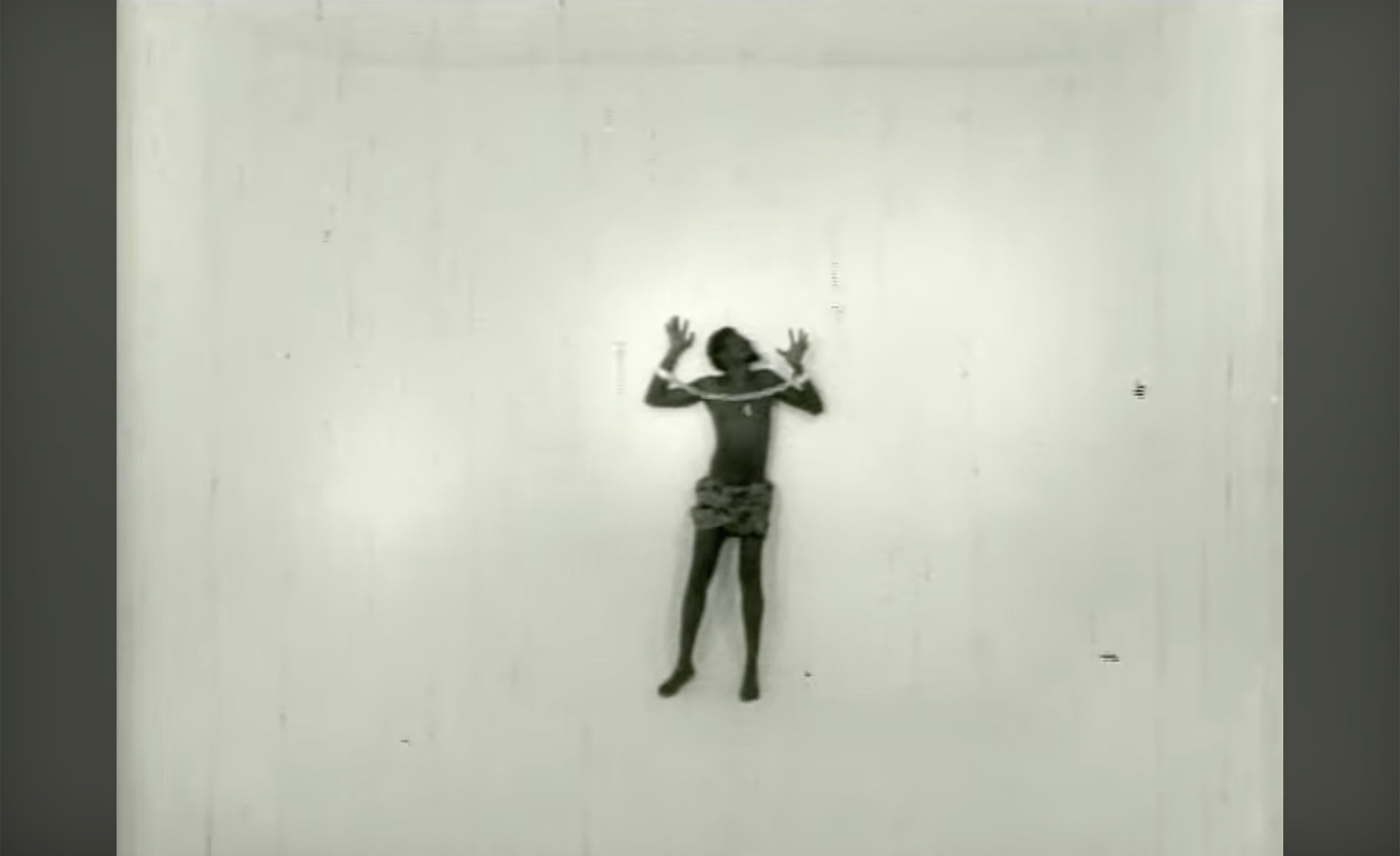 A dark-skinned person, hands raised and bound by ropes across their shoulders, wearing a patterned skirt, against a plain white wall.