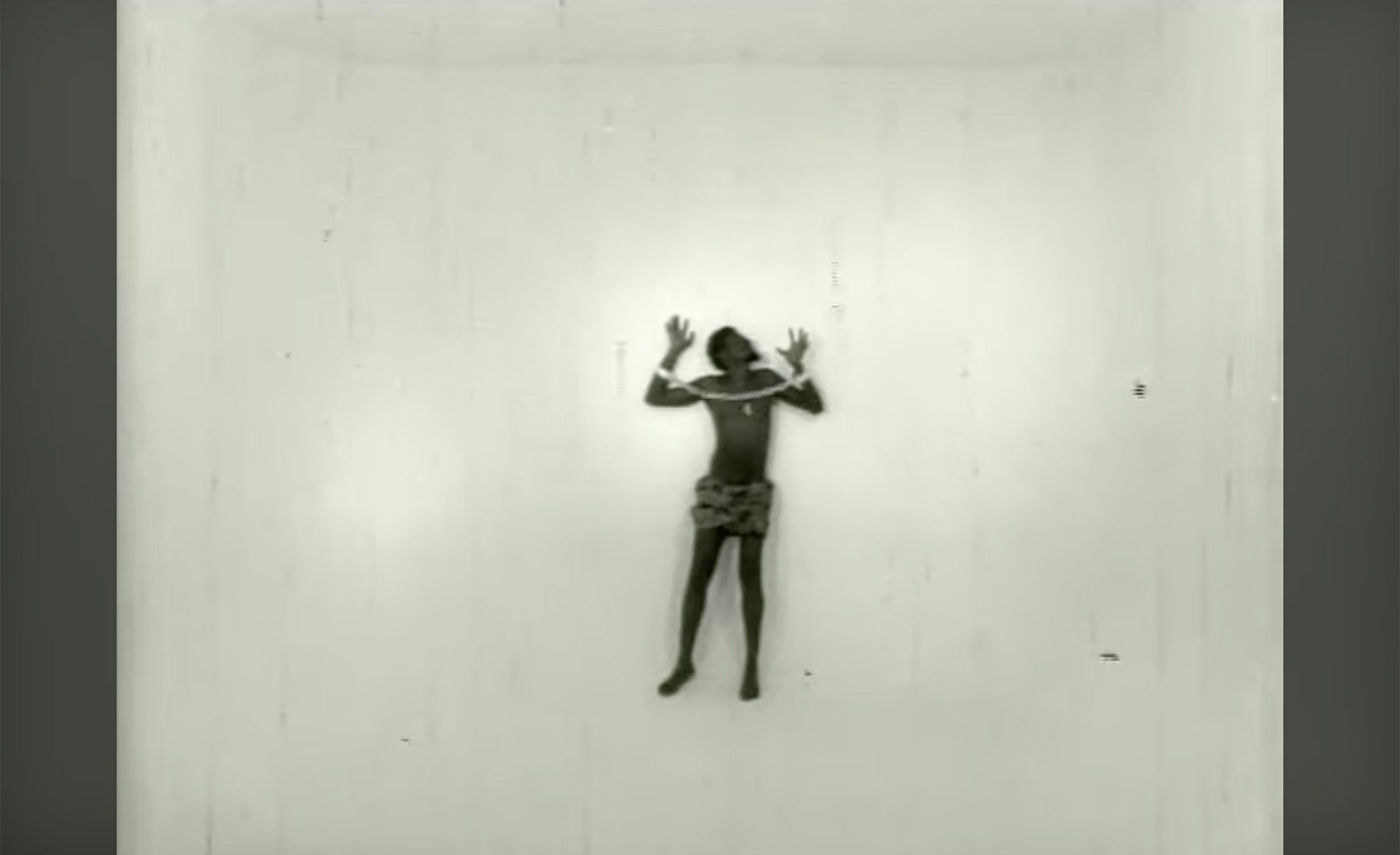 A dark-skinned person, hands raised and bound by ropes across their shoulders, wearing a patterned skirt, against a plain white wall.