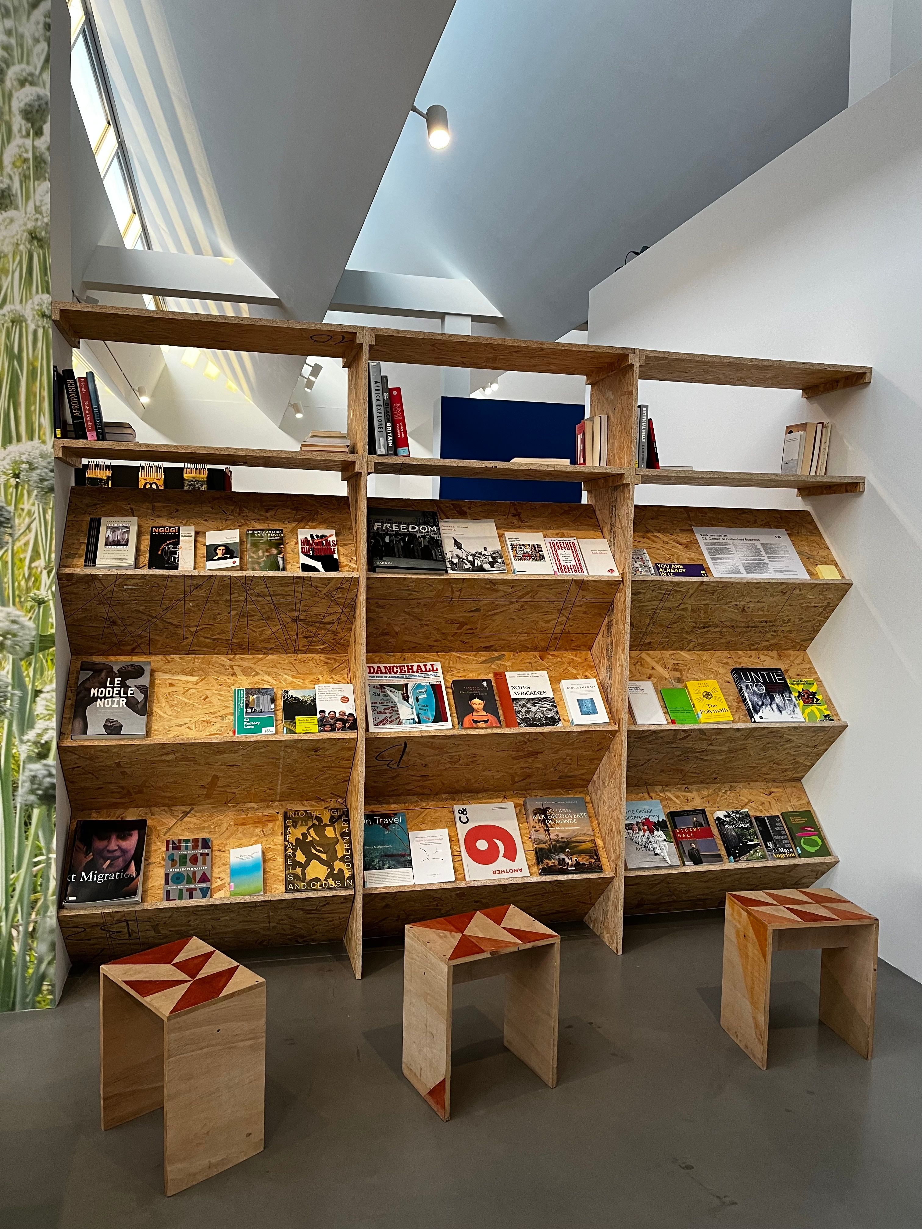A modern bookstore interior with three tall wooden display shelves full of books, and three small wooden stools with red geometric patterns.