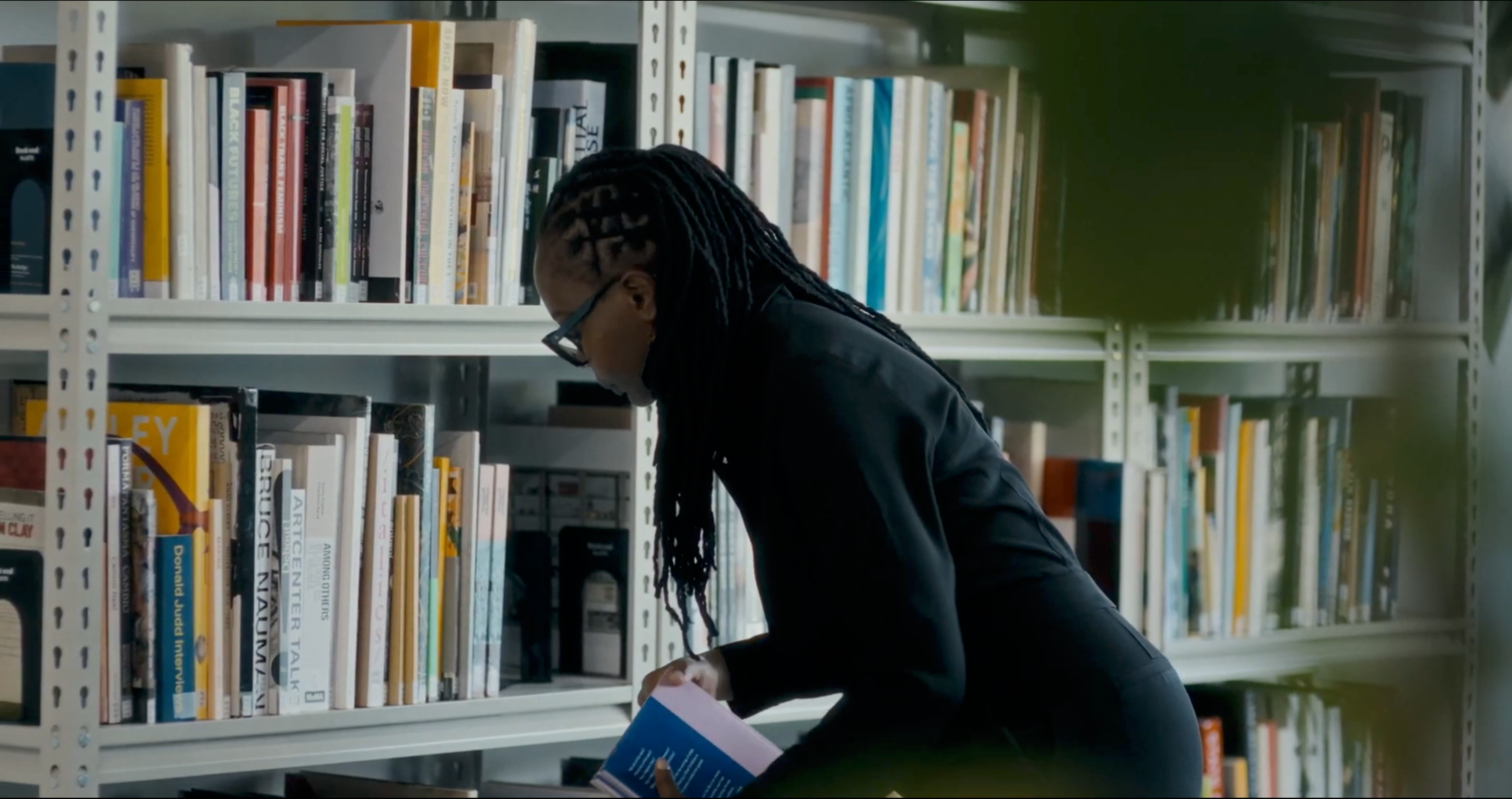 A person with dark braided hair and glasses examines a book in a library.