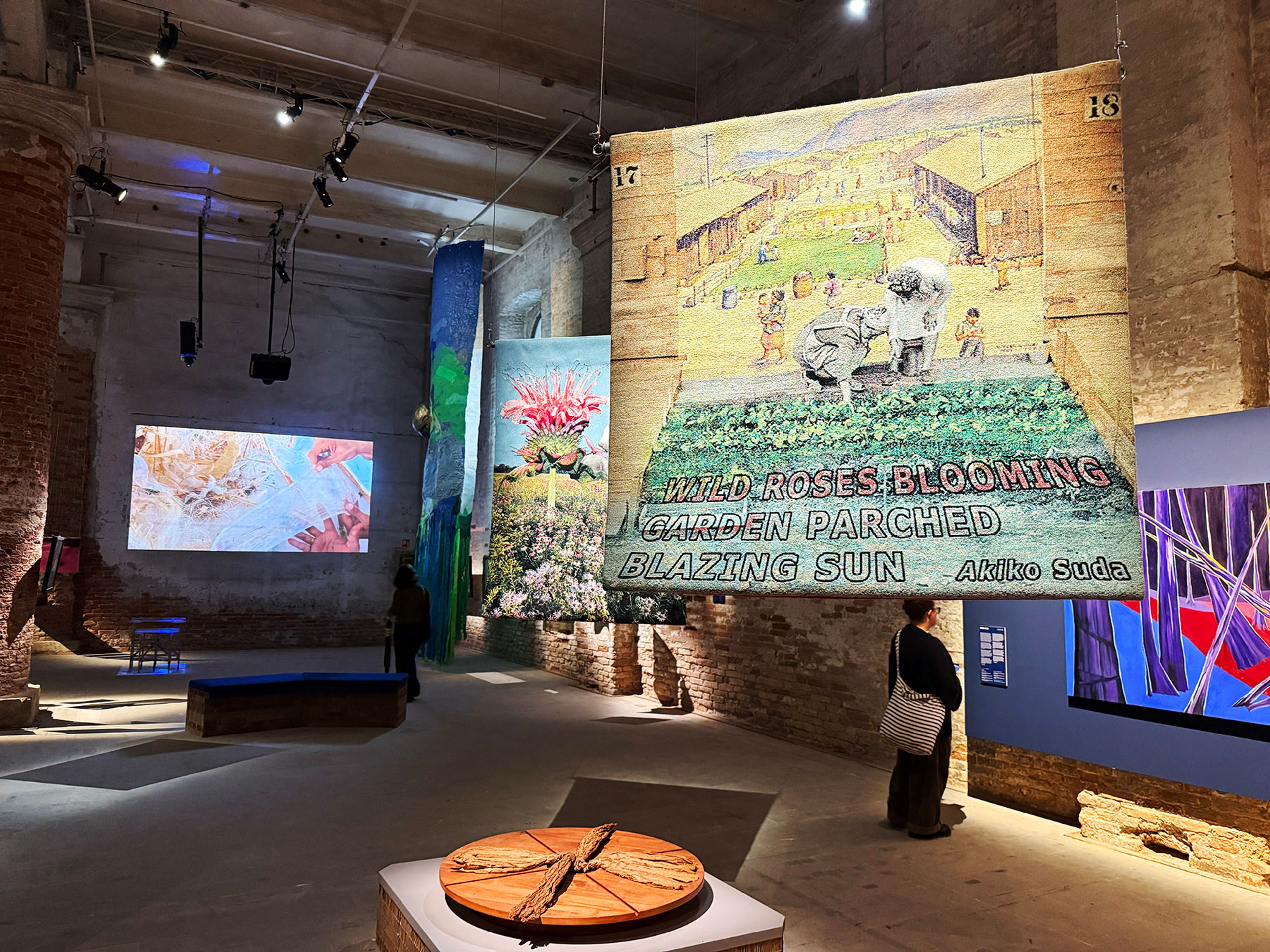 Art exhibition in a high-ceilinged, brick-walled gallery, featuring large hanging tapestries, projected video art, and a person viewing.
