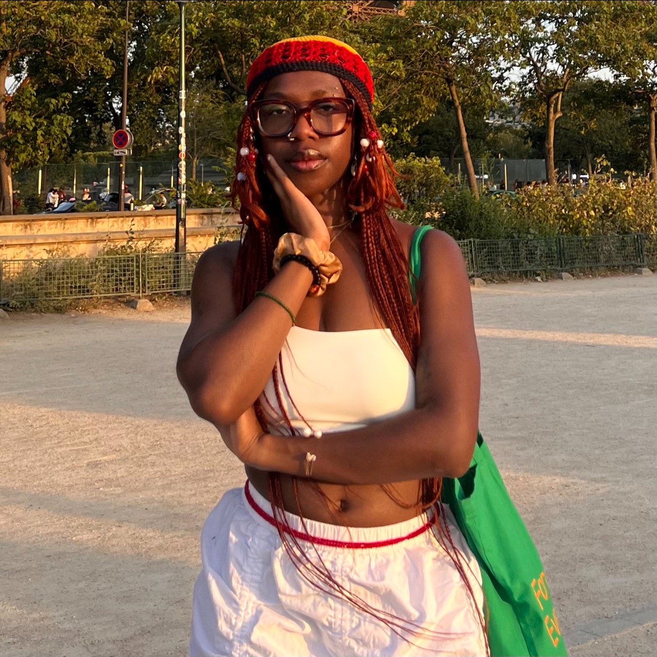 Young Black woman with reddish braids, red-yellow-green beanie, white crop top and pants, poses outdoors, hand on cheek.