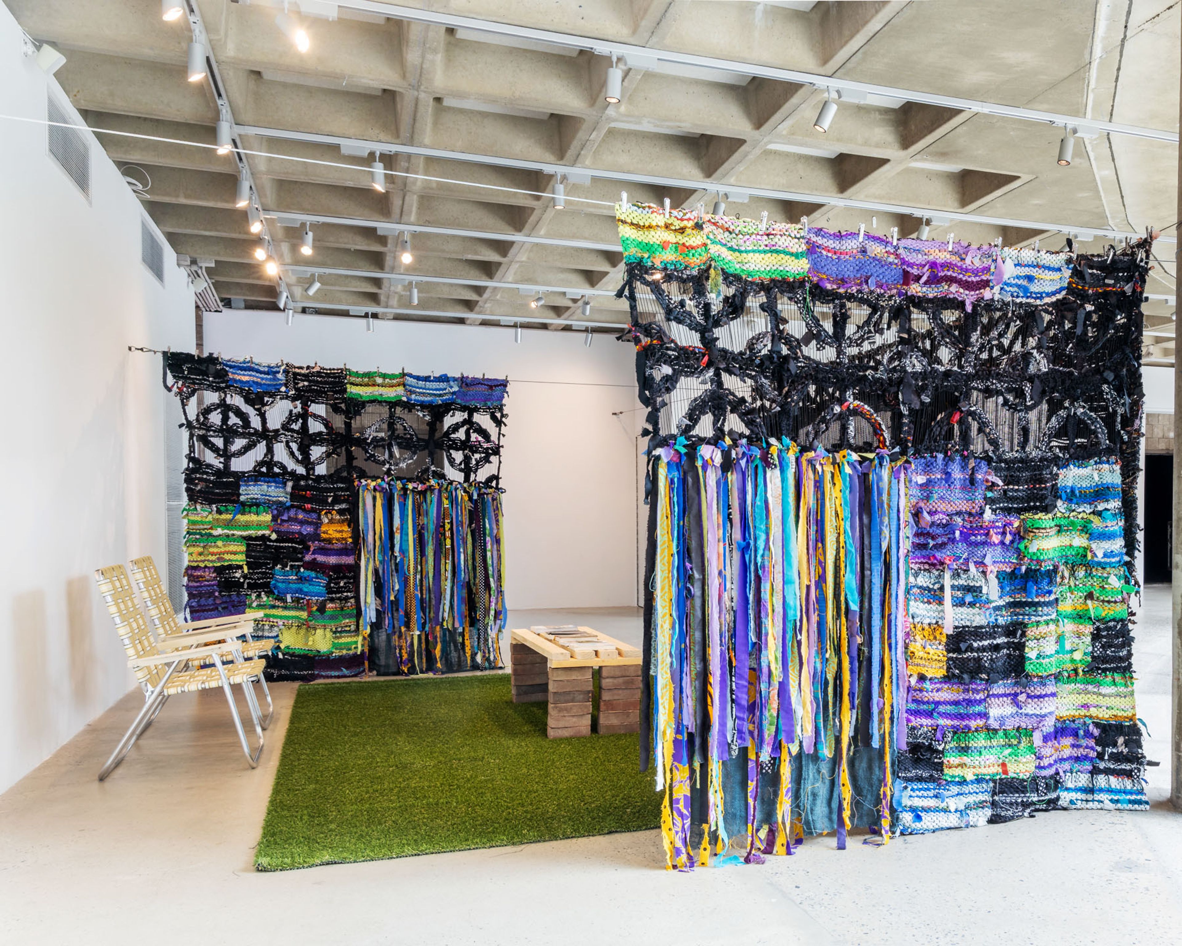 Colorful woven walls with circular patterns and hanging ribbons, a green rug, and chairs in a modern gallery.