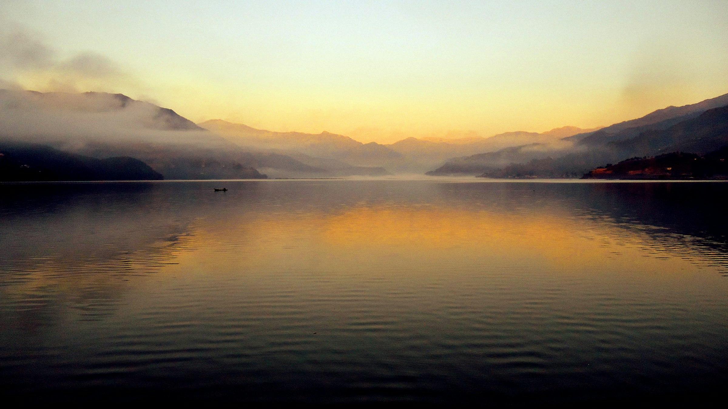 A calm lake reflects a golden dawn sky, with misty hills on both shores and a single small boat on the water.