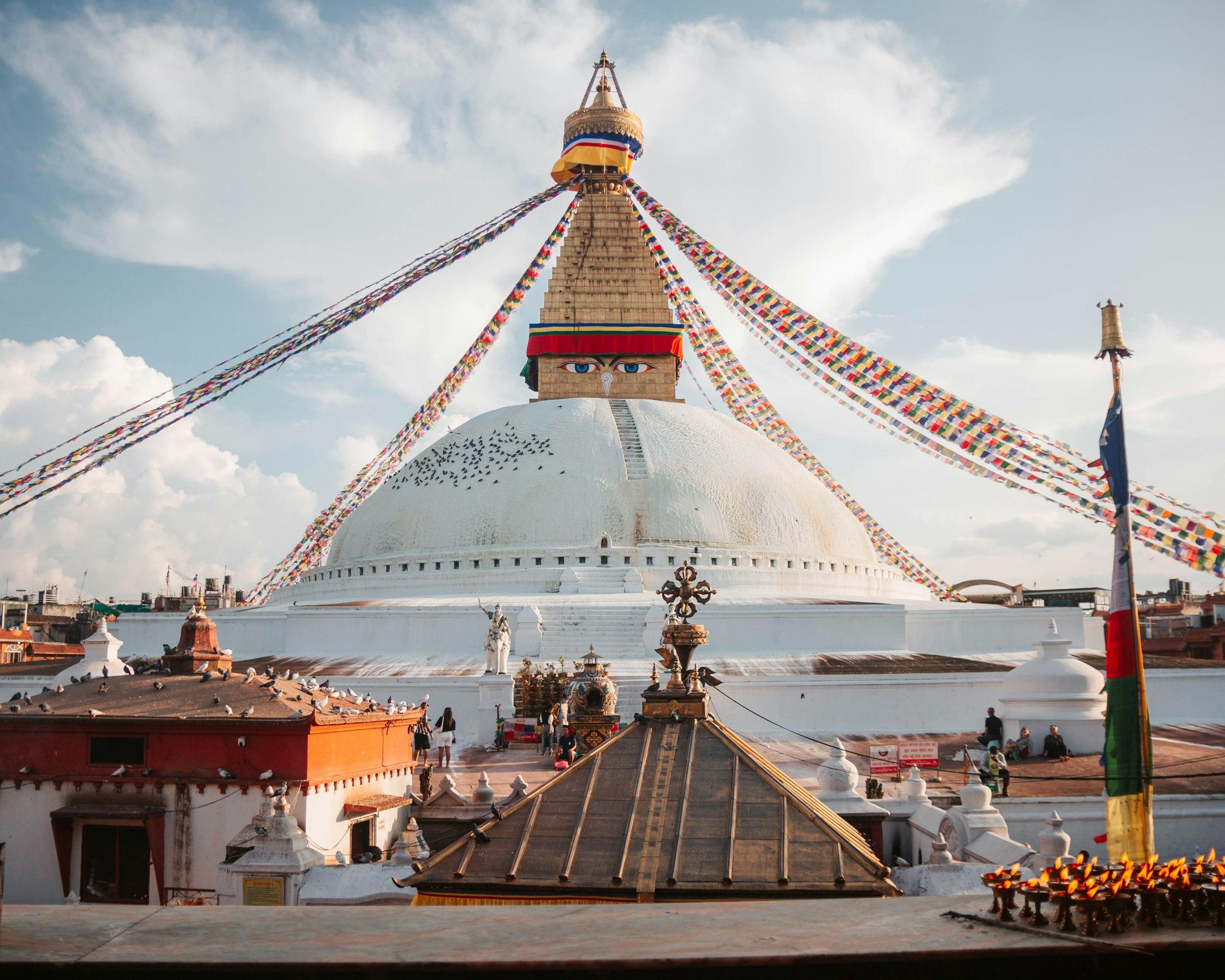 The white dome and gilded spire of Boudhanath Stupa with painted eyes and strings of prayer flags, seen above rooftops and butter lamps.