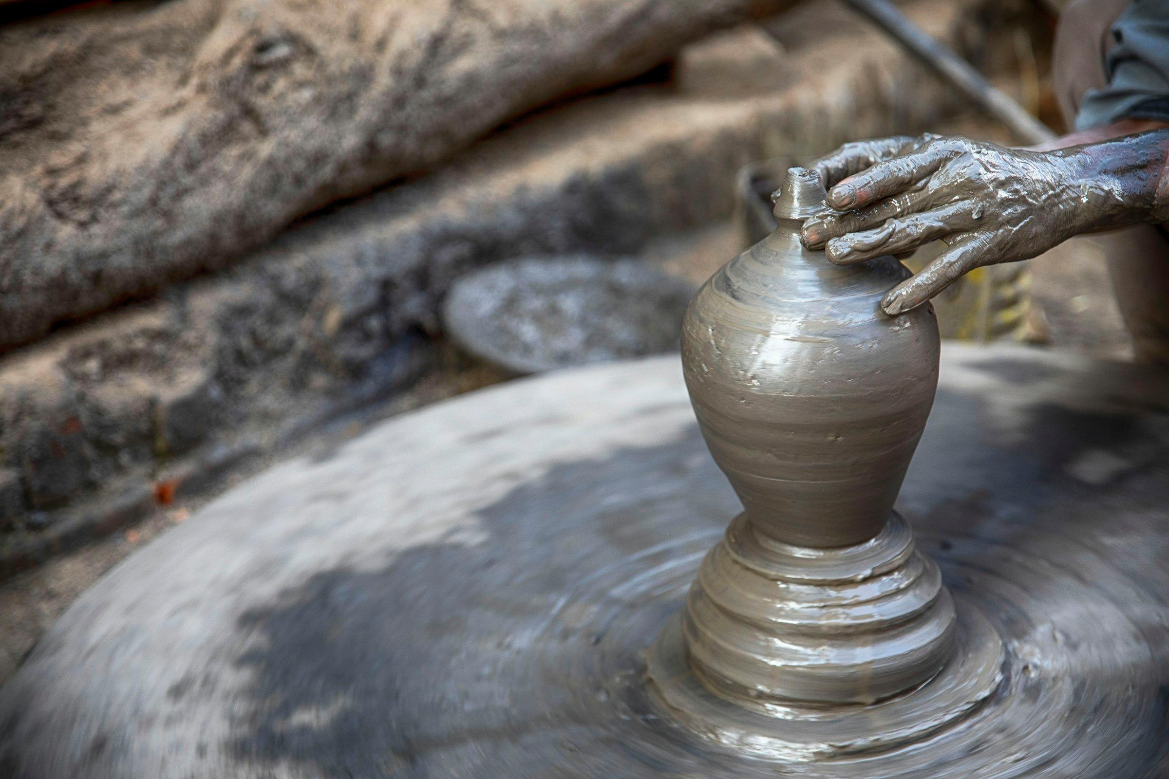 A potter’s clay-covered hand shapes a vase on a spinning wheel; only the hand is visible.