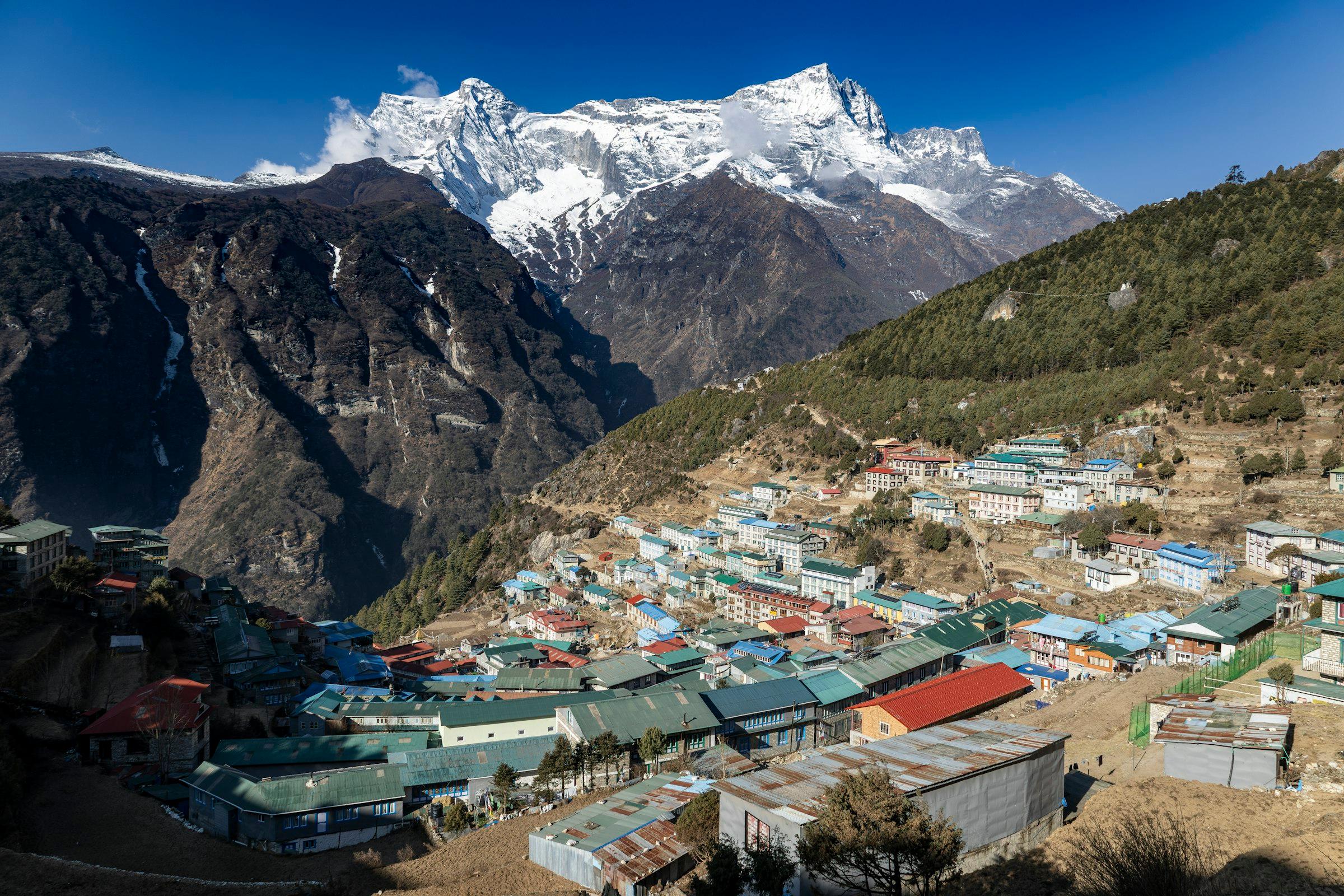 Terraced houses with coloured roofs fill a horseshoe-shaped hillside below a large snow-covered mountain.