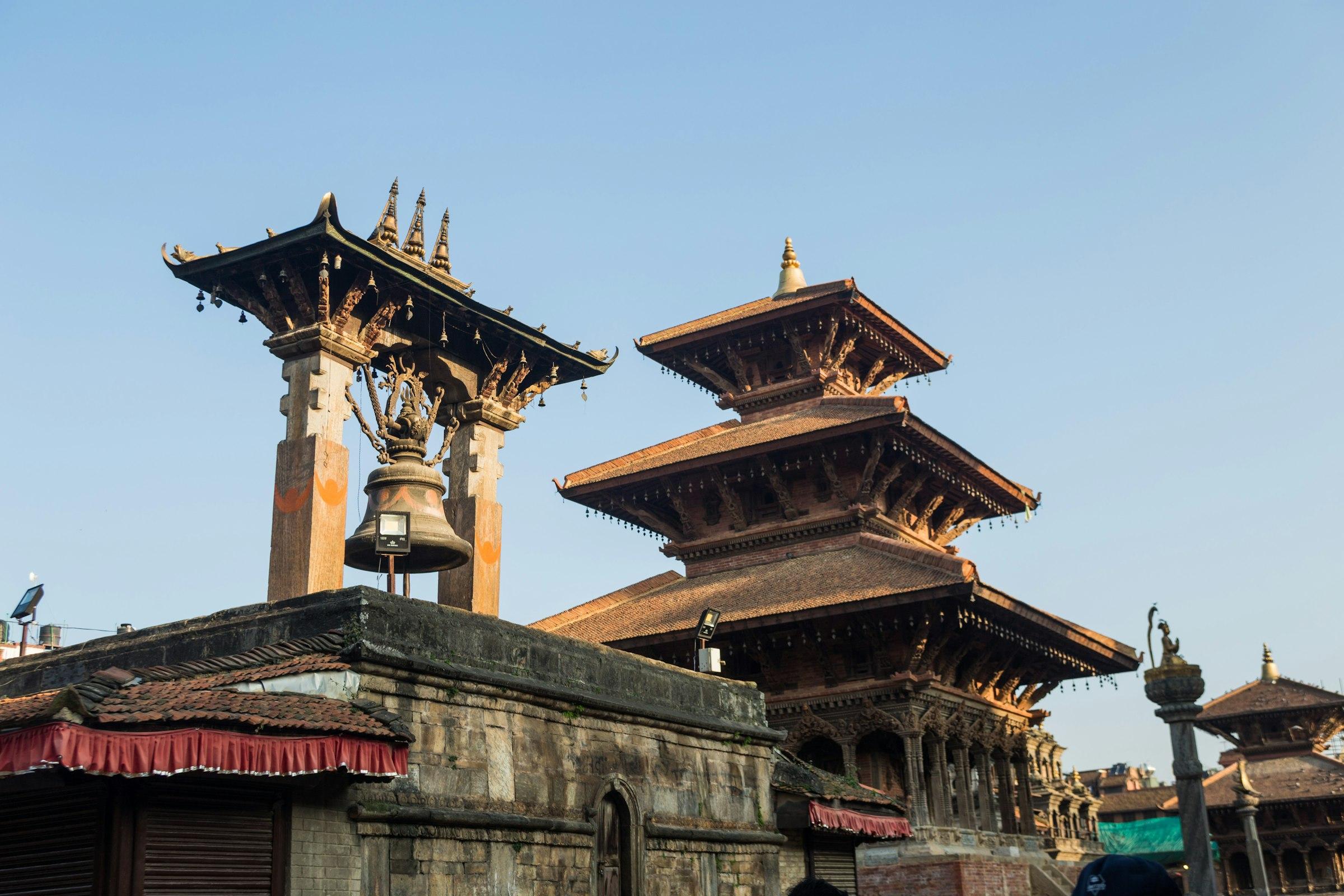 A large bronze bell in a carved pavilion beside a three-tiered pagoda temple against a clear sky in warm light.