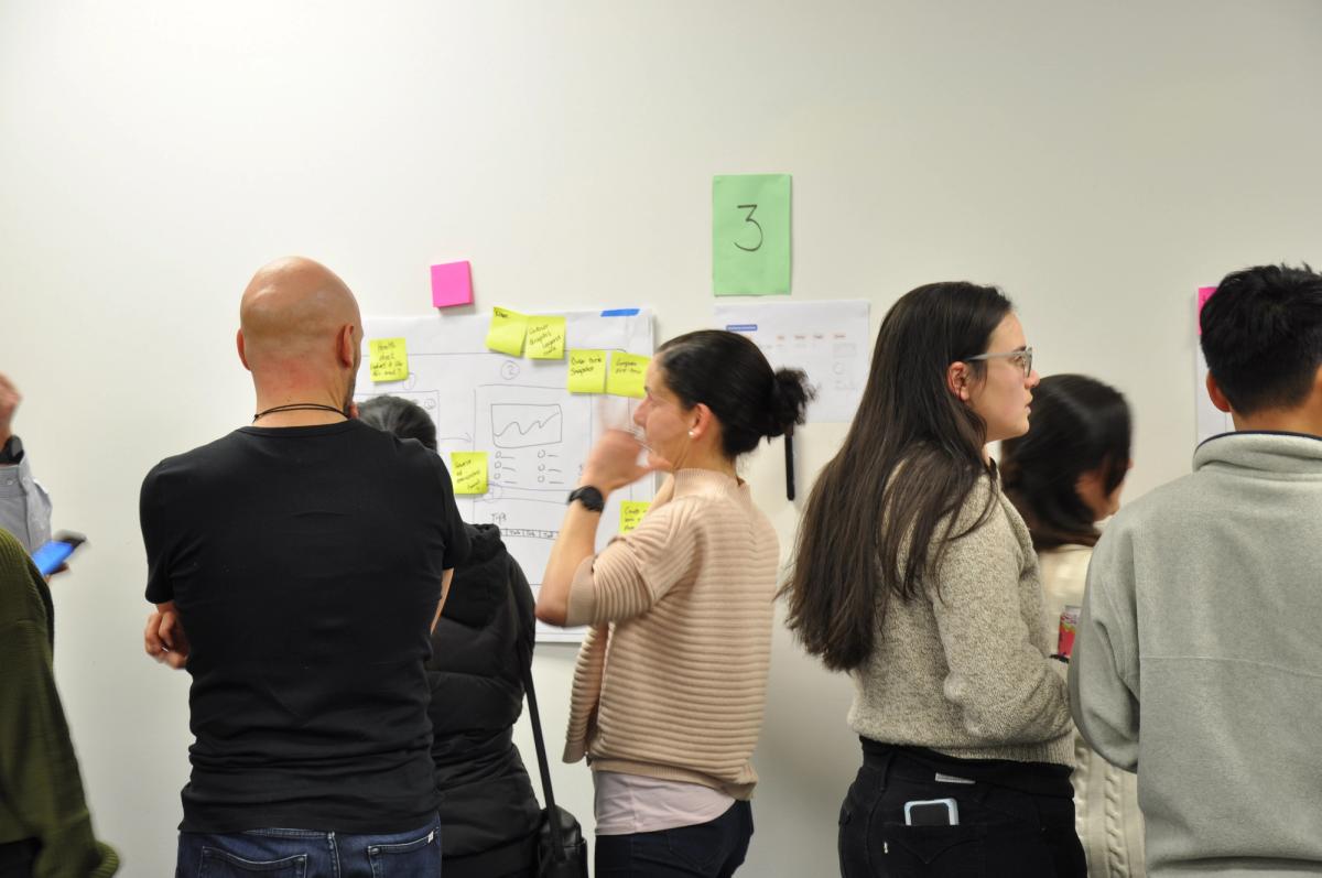 A group of people talking with a poster of a wireframe and post it notes on the wall