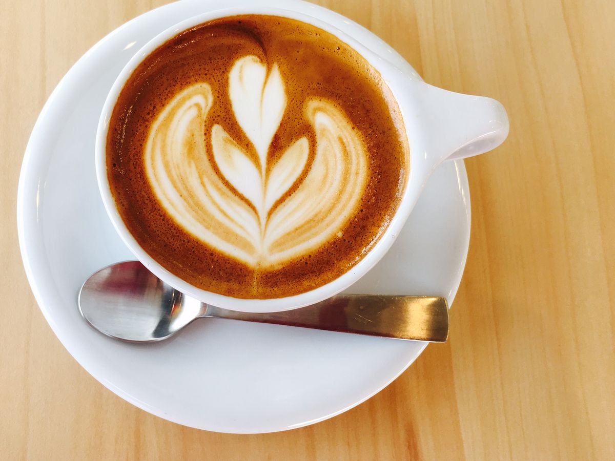 A flat white in a white cup on a white saucer with a teaspoon on the saucer