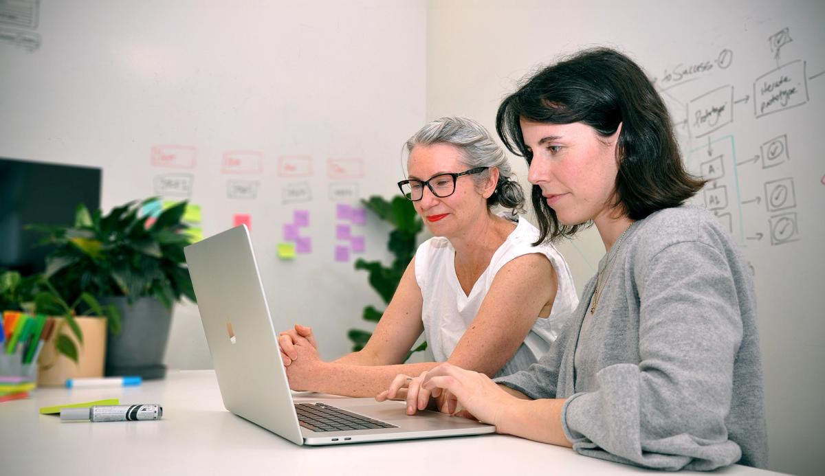 Two people looking at a laptop with post-it notes and drawings on the wall behind.