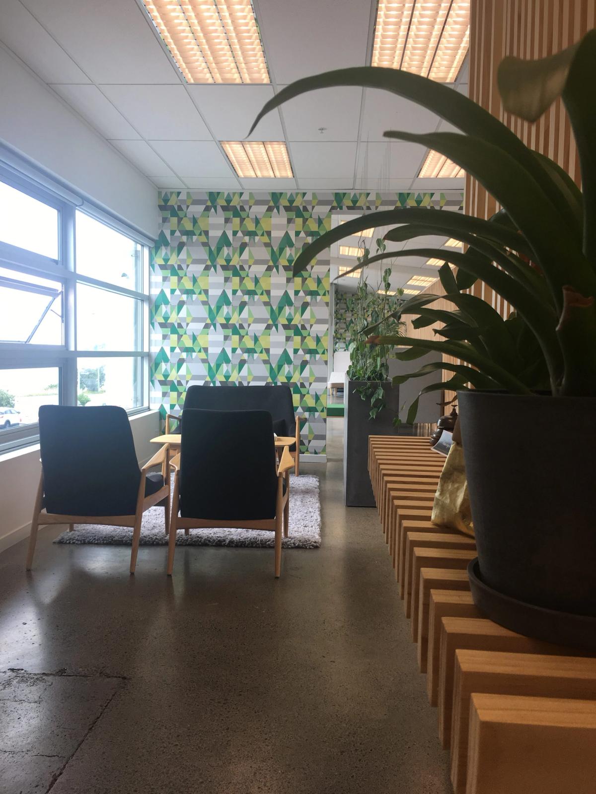 Seating area bordered by windows on the left and a wall in the background. Plants in the foreground. 