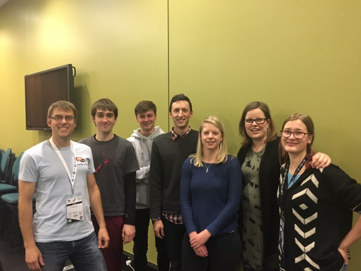 Group of 7 people standing if front of an olive green wall