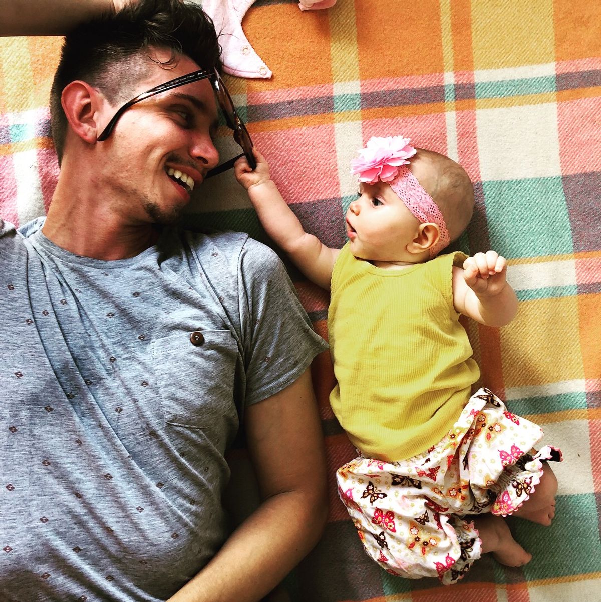 Dad and baby lying on a blanket looking at each other, baby is pulling sunglasses off dad's face
